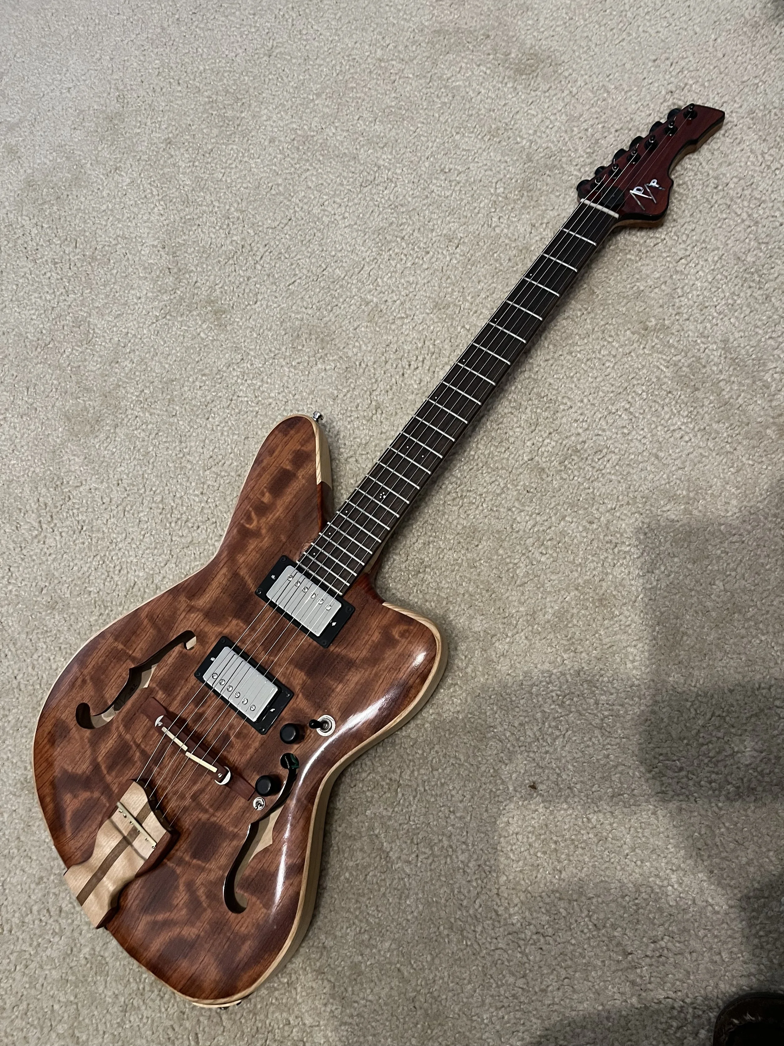 A semi-hollow electric guitar with a natural wood finish and black hardware laying on a beige carpeted floor.