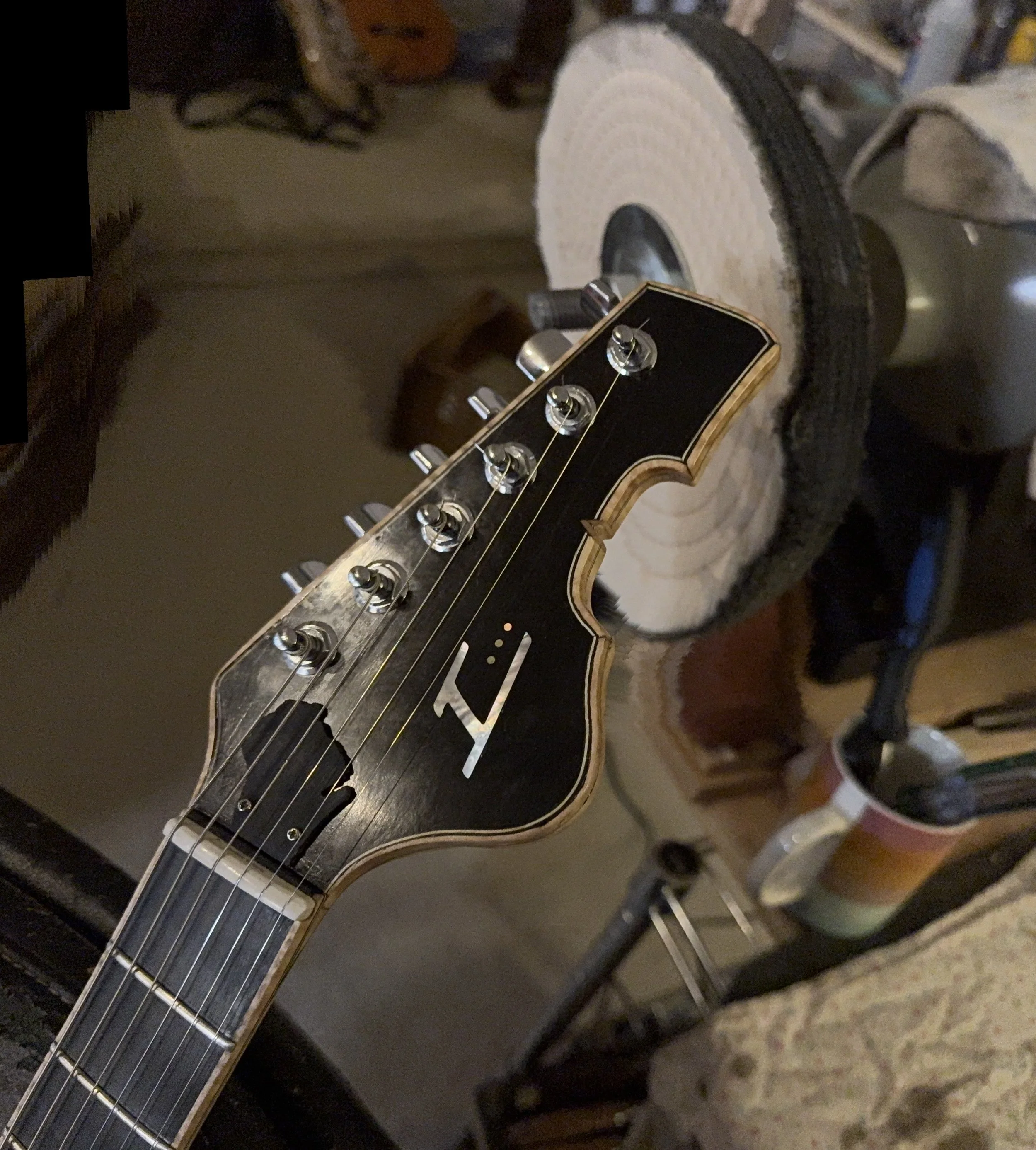 Close-up of an electric guitar's headstock with tuning pegs, strings, and truss rod cover, resting in a room with various items in the background.