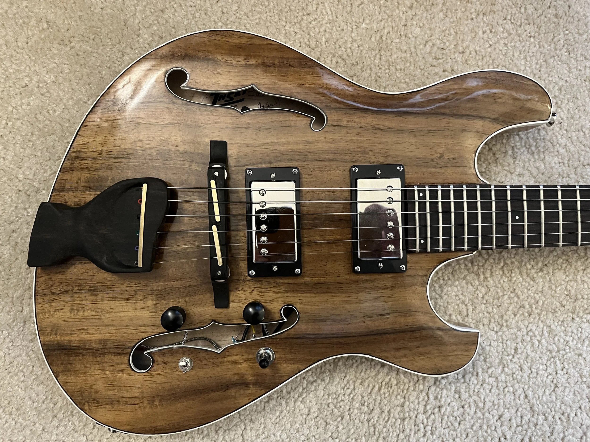 Close-up of a wooden electric guitar body with black and white binding, two humbucker pickups, a bridge, and control knobs, lying on a beige carpet.