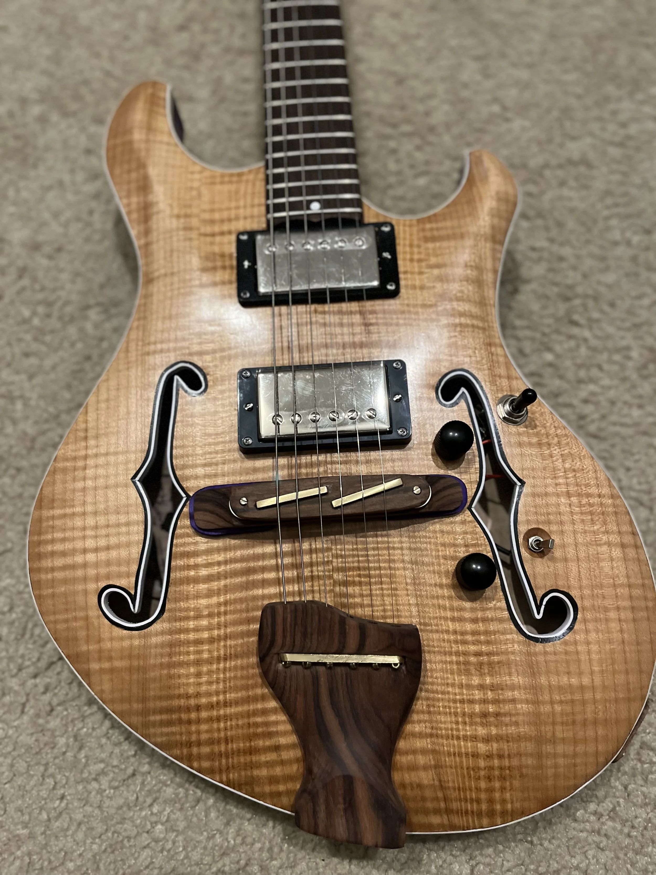 A semi-hollow electric guitar with a natural wood finish, f-holes, two pickups, and a dark wooden pickguard, placed on a beige carpet.