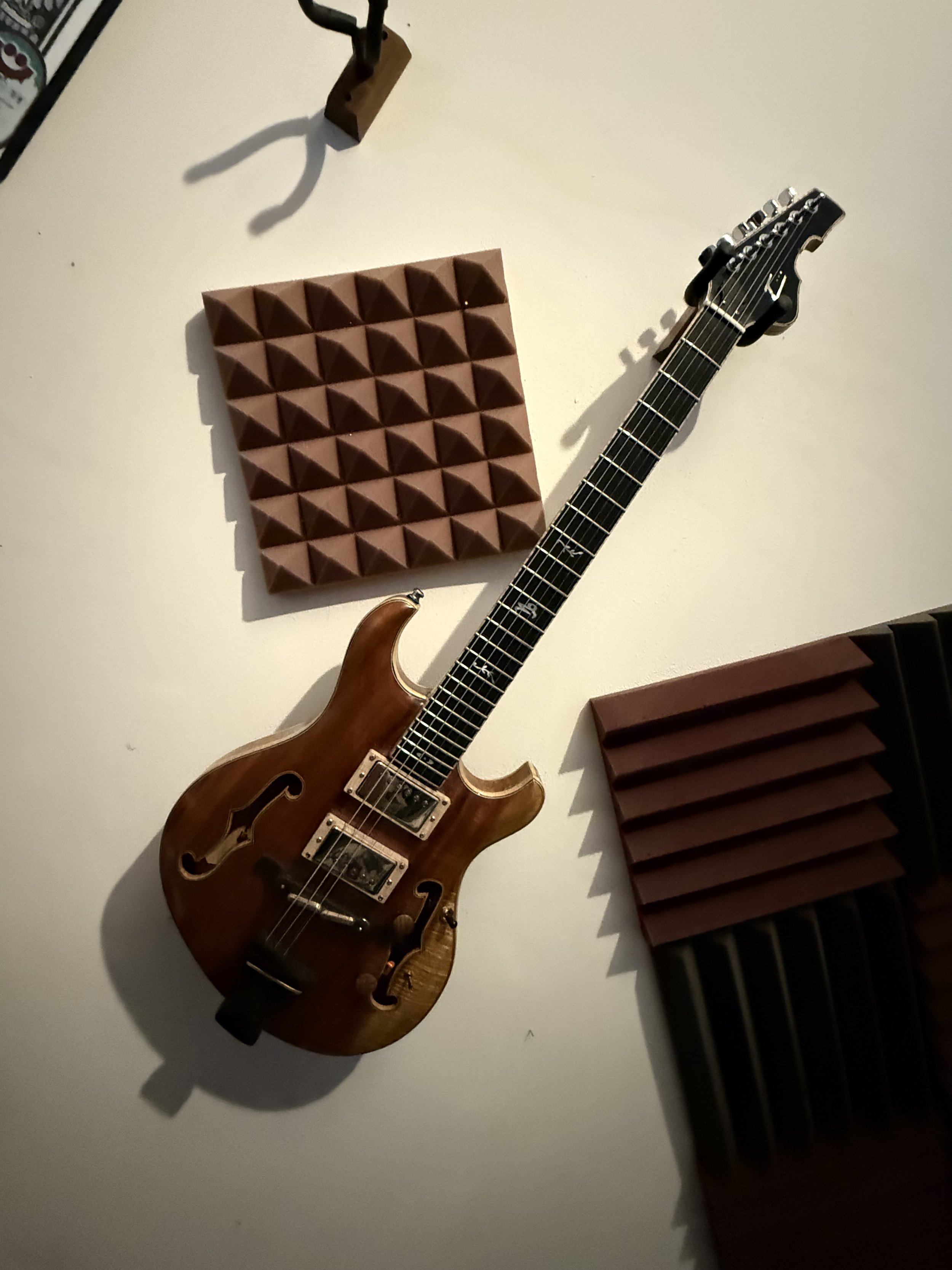 A semi-hollow electric guitar with a wooden finish hanging on a white wall, accompanied by acoustic foam panels for soundproofing.