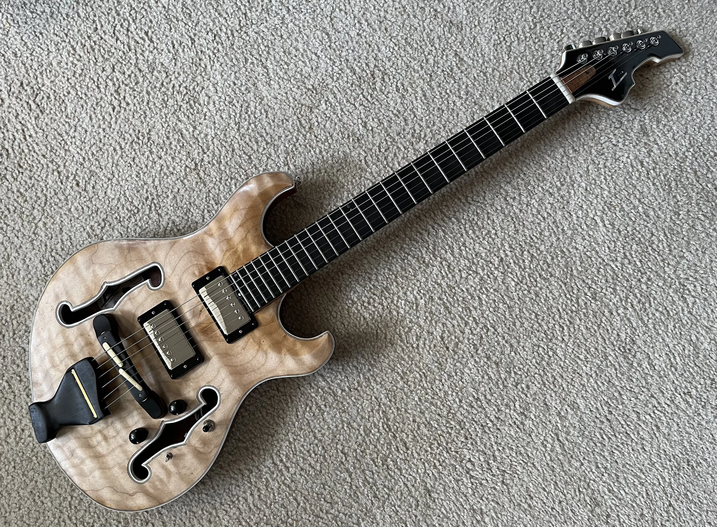 A semi-hollow electric guitar with a natural wood finish, black hardware, and a black pickguard, lying on a beige textured carpet.