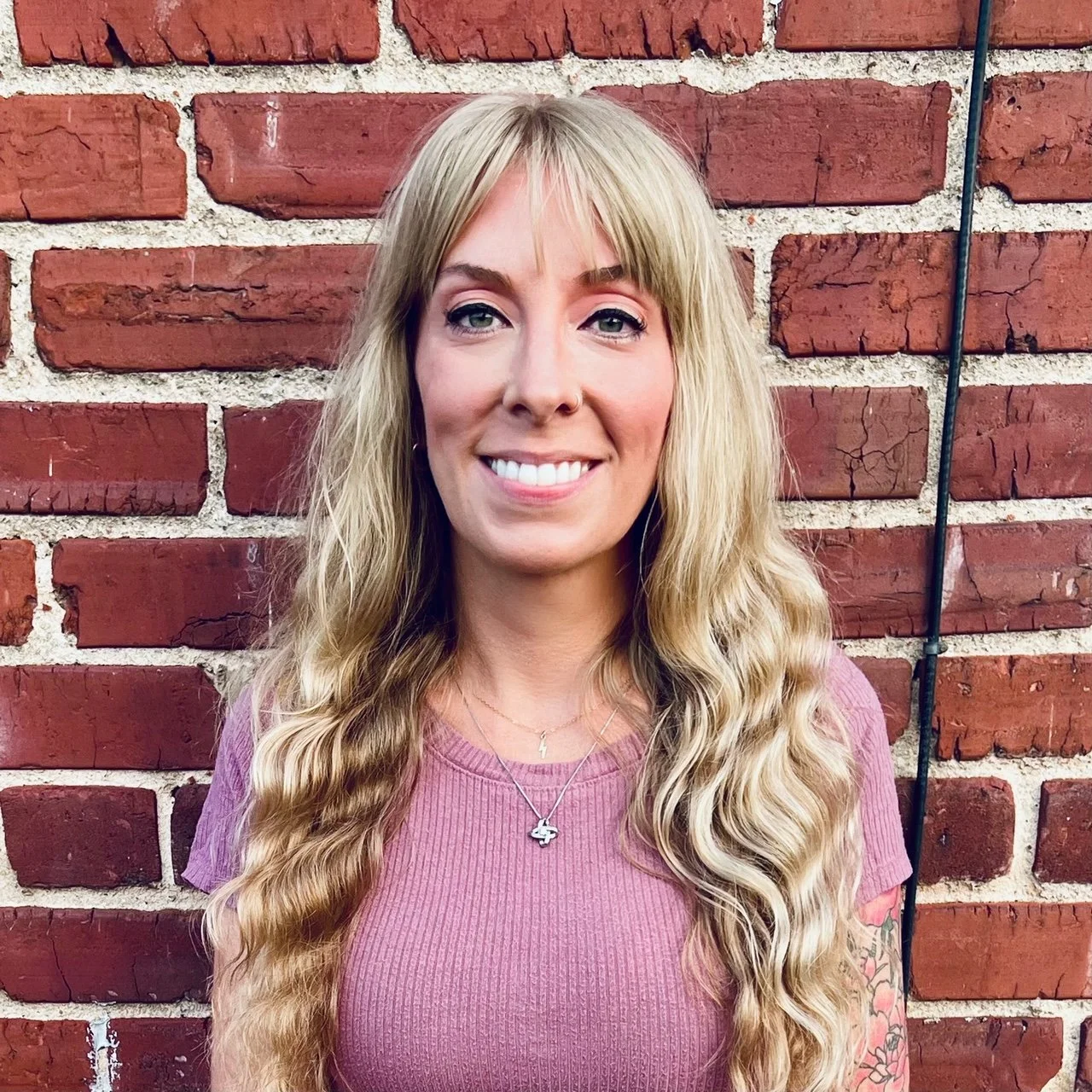 A woman with long, wavy blonde hair, wearing a pink top, smiling, standing in front of a red brick wall.
