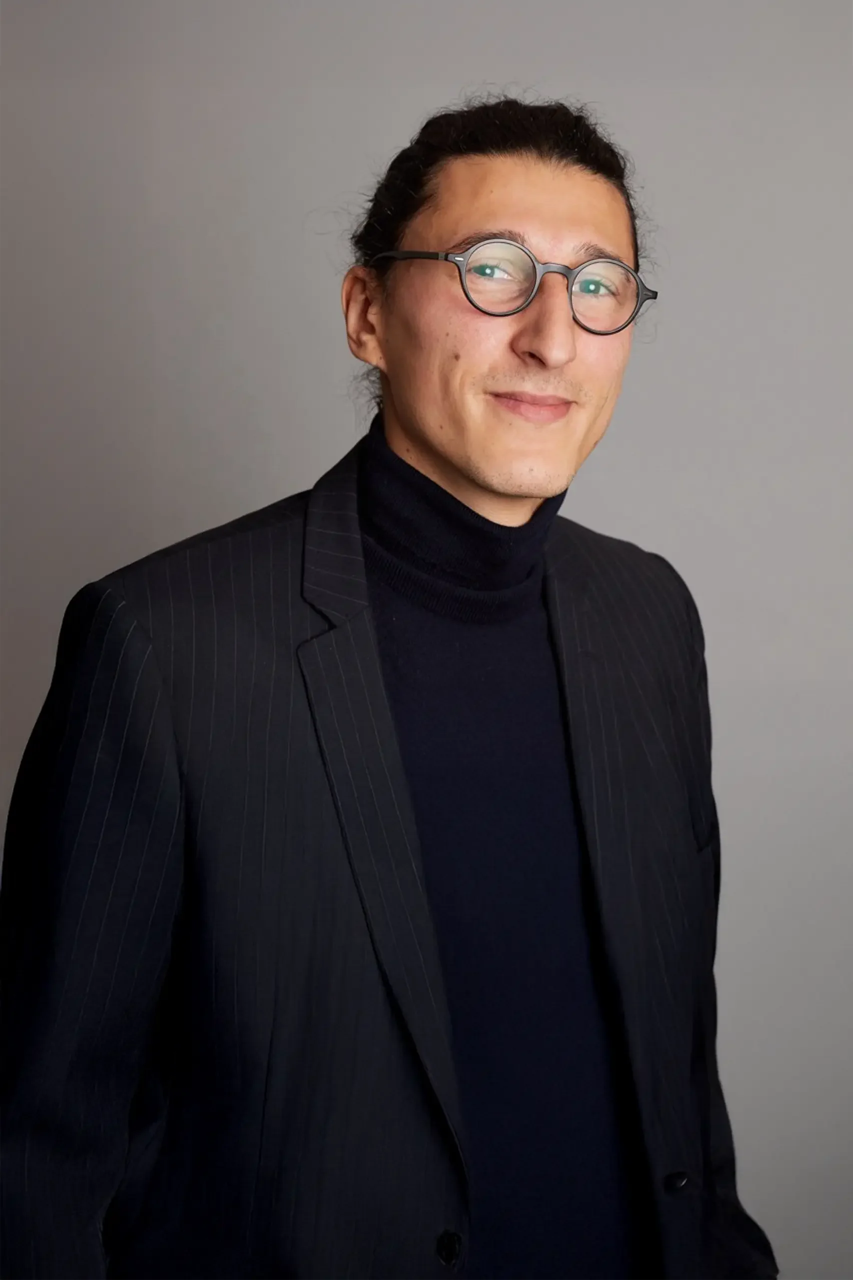 Portrait d'un homme avec des lunettes rondes, portant un costume noir à fines rayures et un turtleneck noir, posé contre un fond gris.