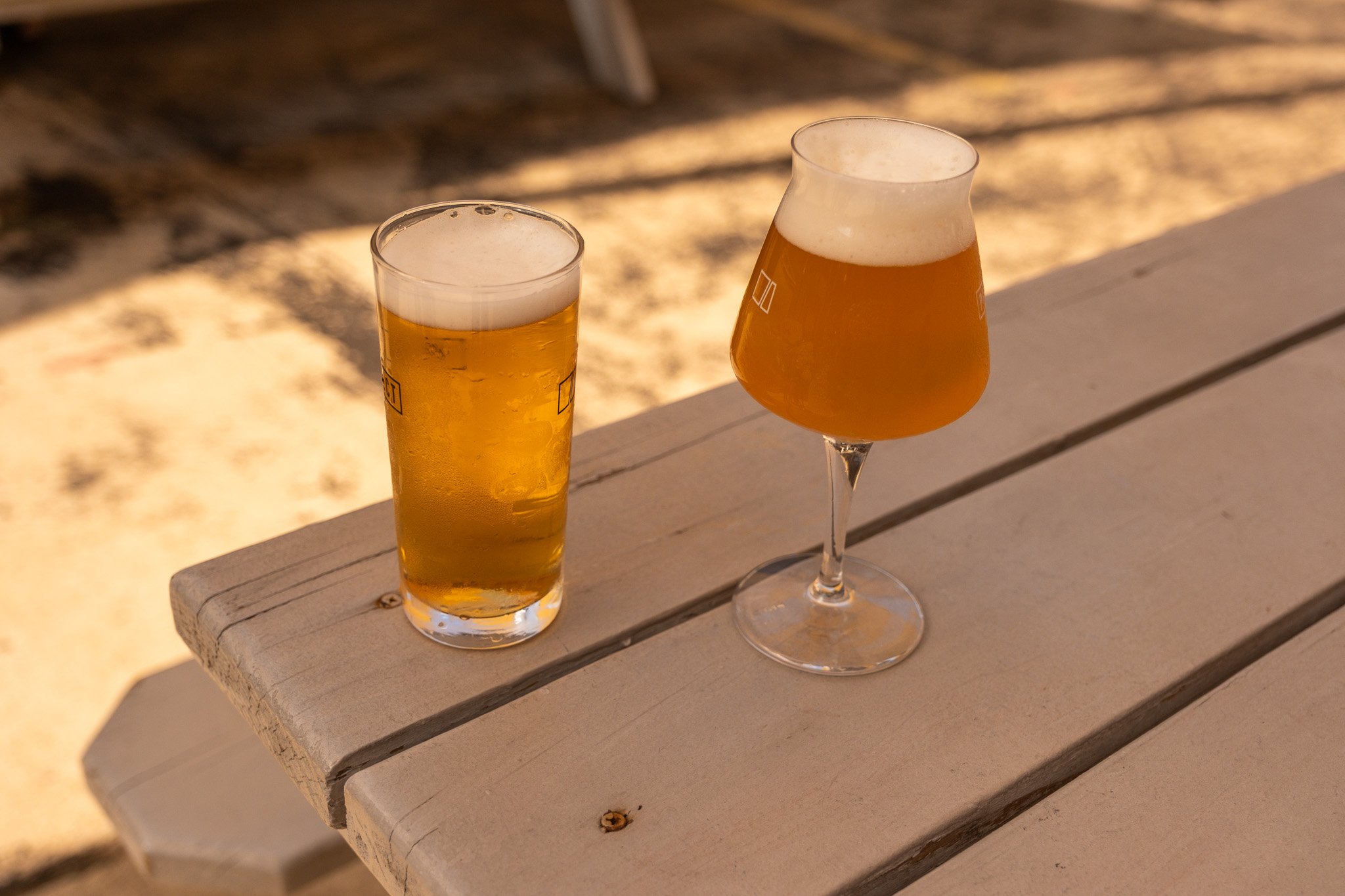 Two glasses of beer on a wooden table outdoors, one in a tall pint glass and the other in a stemmed glass, both topped with foam.