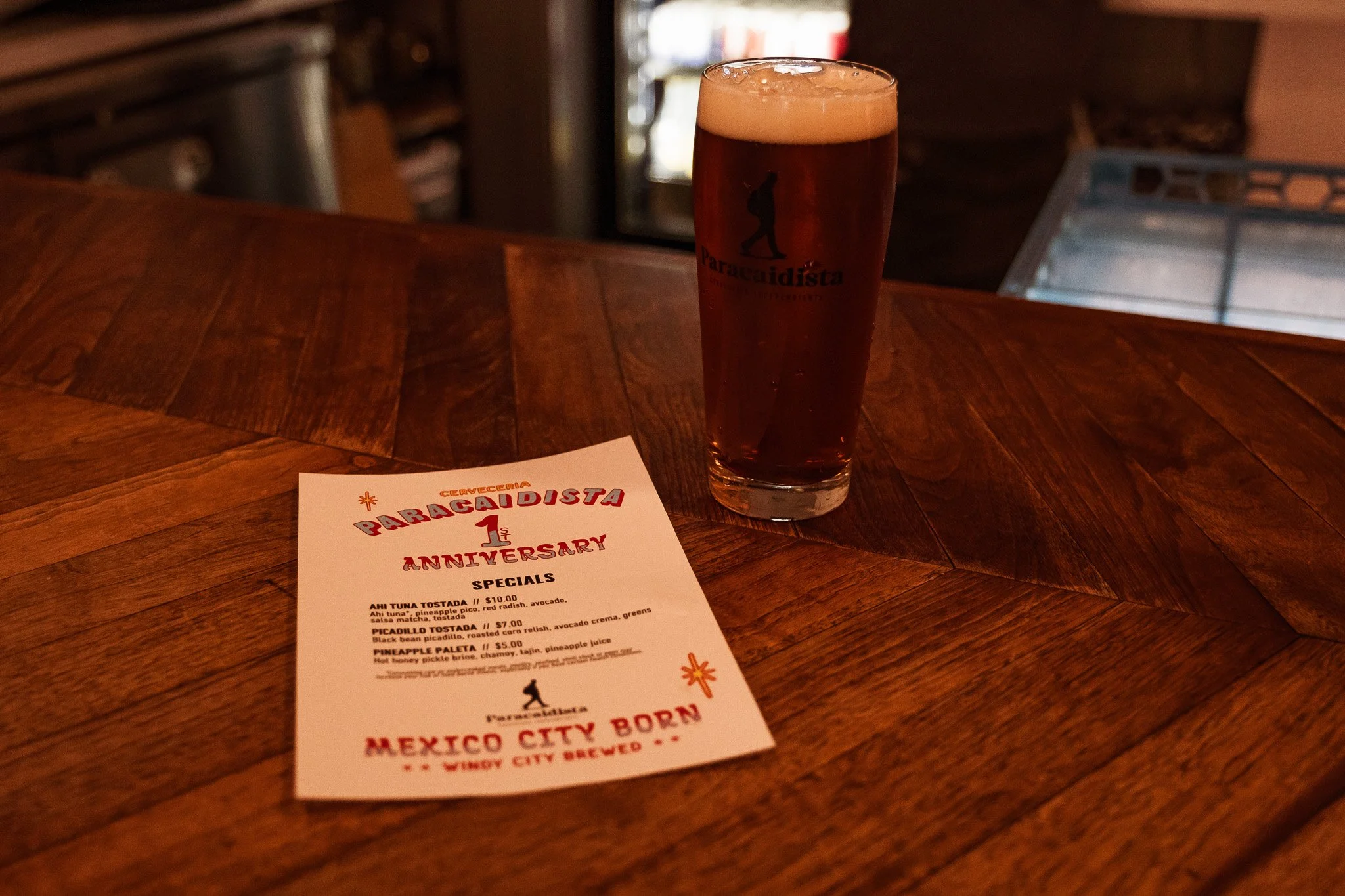 A pint glass filled with dark beer with a light foam head, placed on a wooden bar counter. The glass has the logo and name of Paracaidista. A printed menu or flyer for Paracaidista's 1st anniversary celebration is also on the bar.