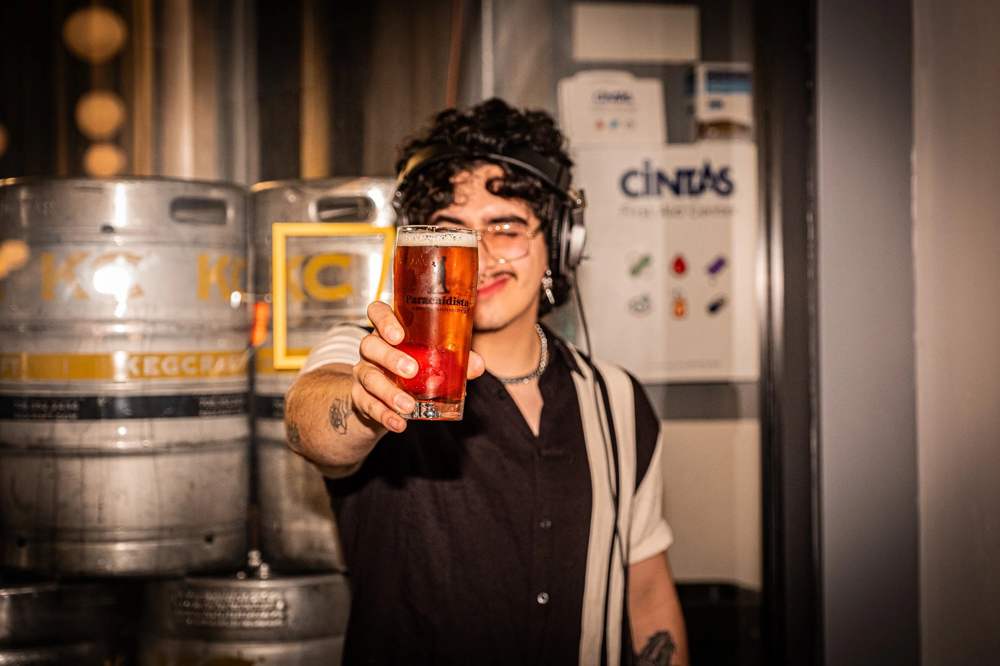 A person with curly hair, glasses, and tattoos smiling and holding a glass of beer towards the camera inside a brewery or pub.