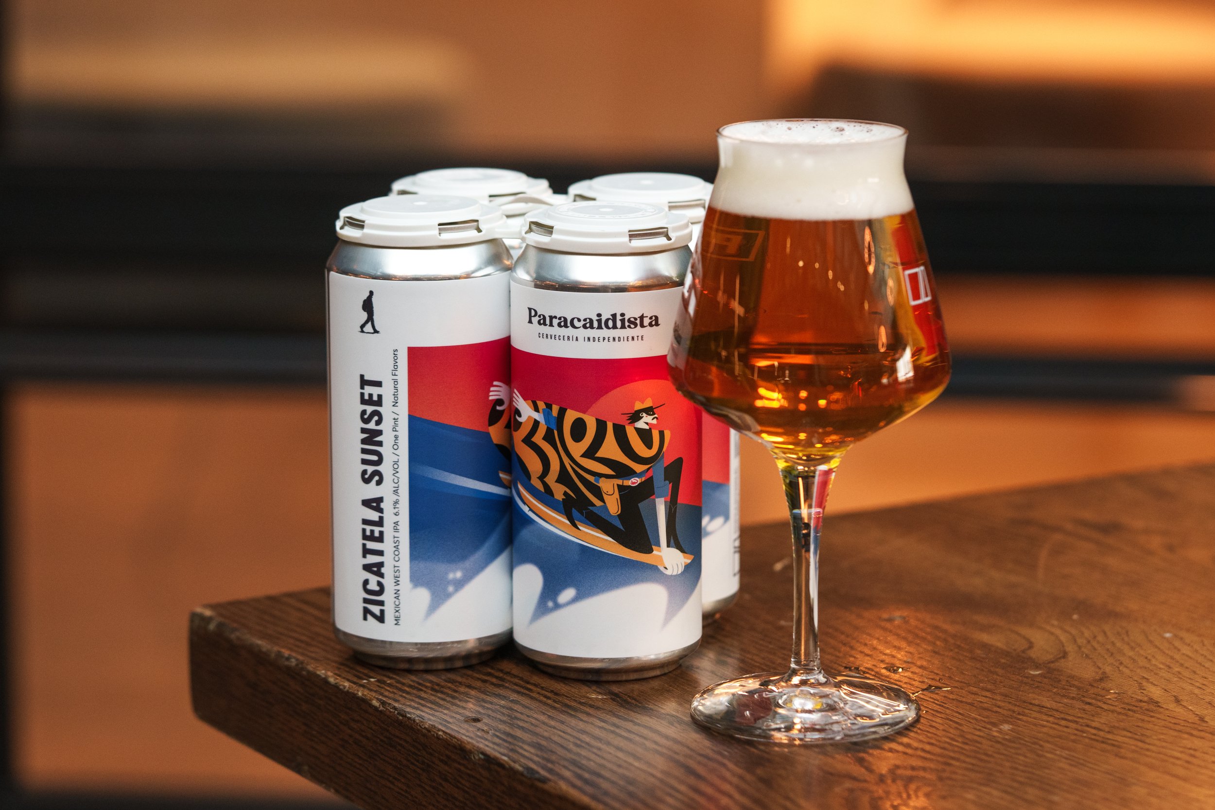 A glass of beer and four cans of Zicatel a Sunset beer on a wooden table.