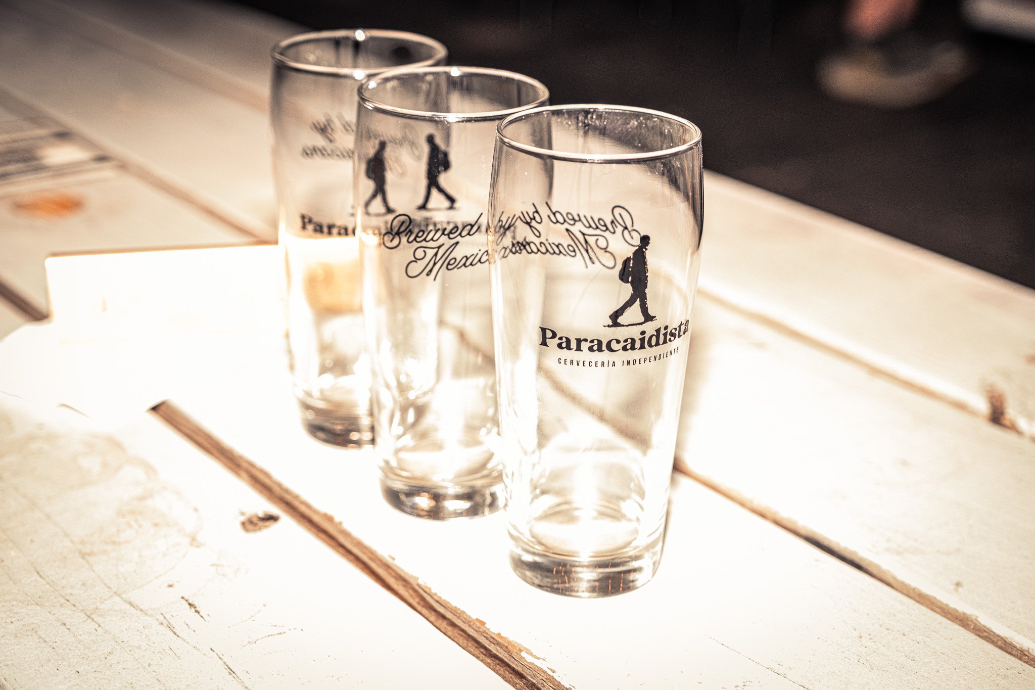 Three clear beer glasses with black logos and text on a white wooden surface, with a blurred background.