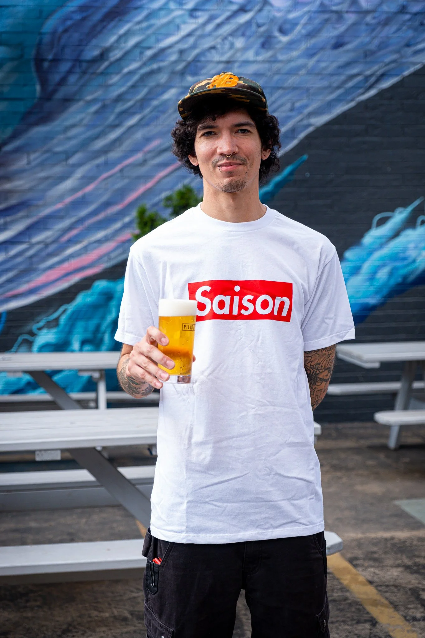 A young man with tattoos, curly hair, and a nose ring is standing outdoors holding a pint of beer, wearing a white t-shirt with 'Saison' printed on it in red, in front of a colorful mural.