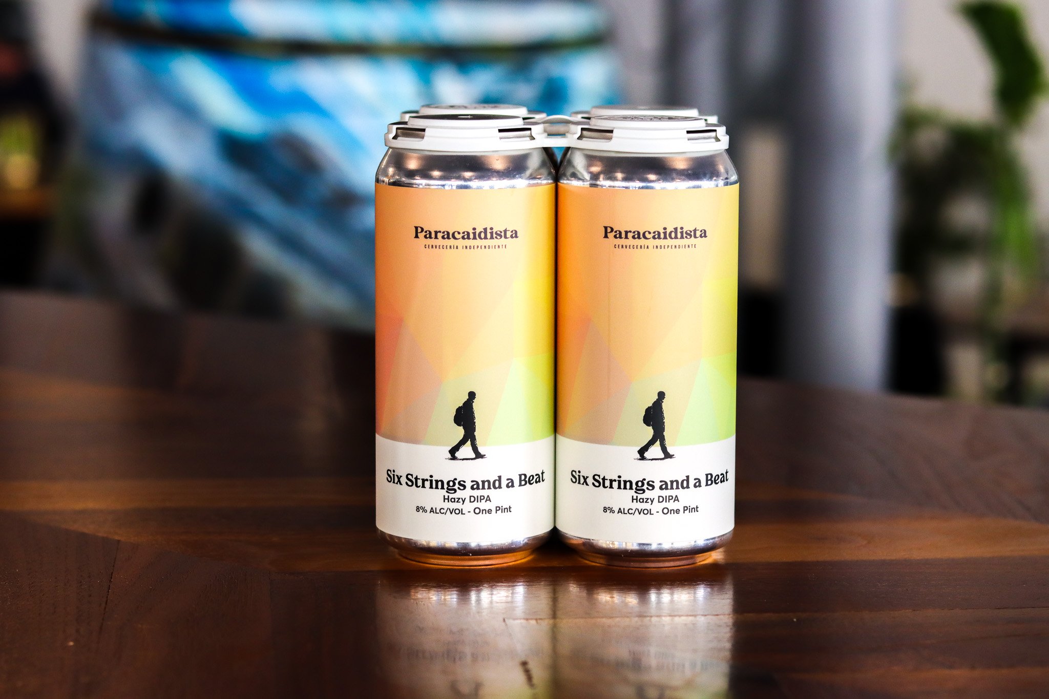 Two aluminum cans of Paracaidista Six Strings and a Beat Hazy DIPA craft beer on a wooden surface, with a blurred background.