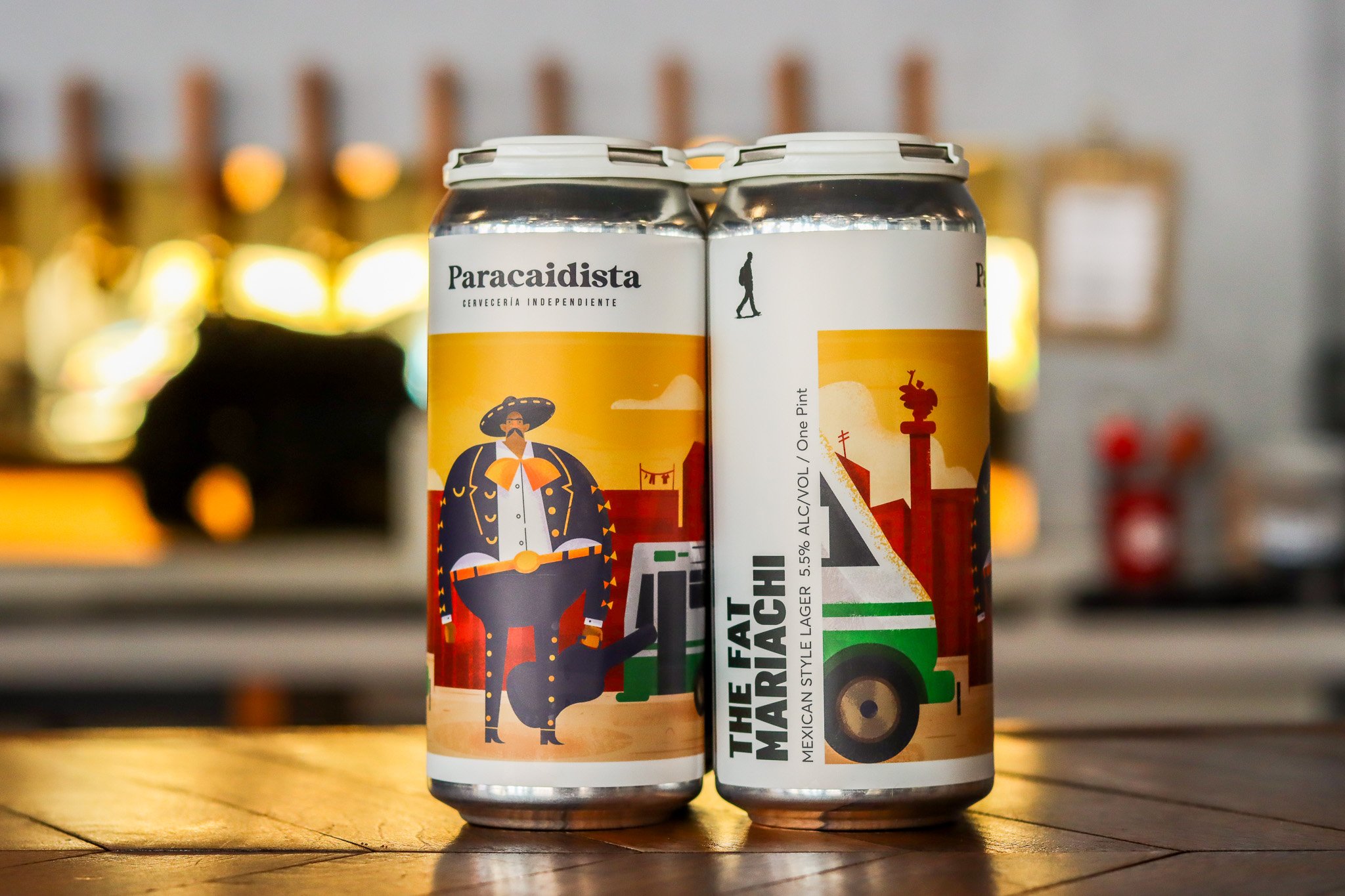 Two cans of beer with colorful artwork, one featuring a man in a sombrero and traditional Mexican clothing, and the other with a Mexican street scene, placed on a wooden surface with a blurred bar background.