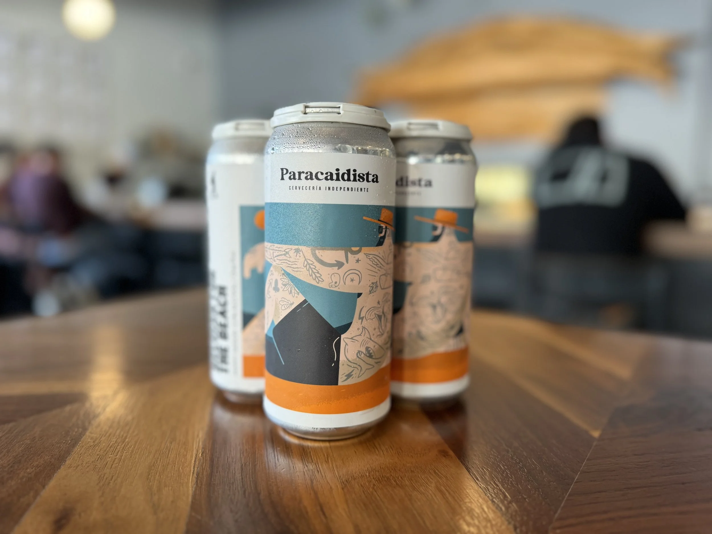 Three cans of Paracaidista craft beer on a wooden table in a restaurant with people in the background.