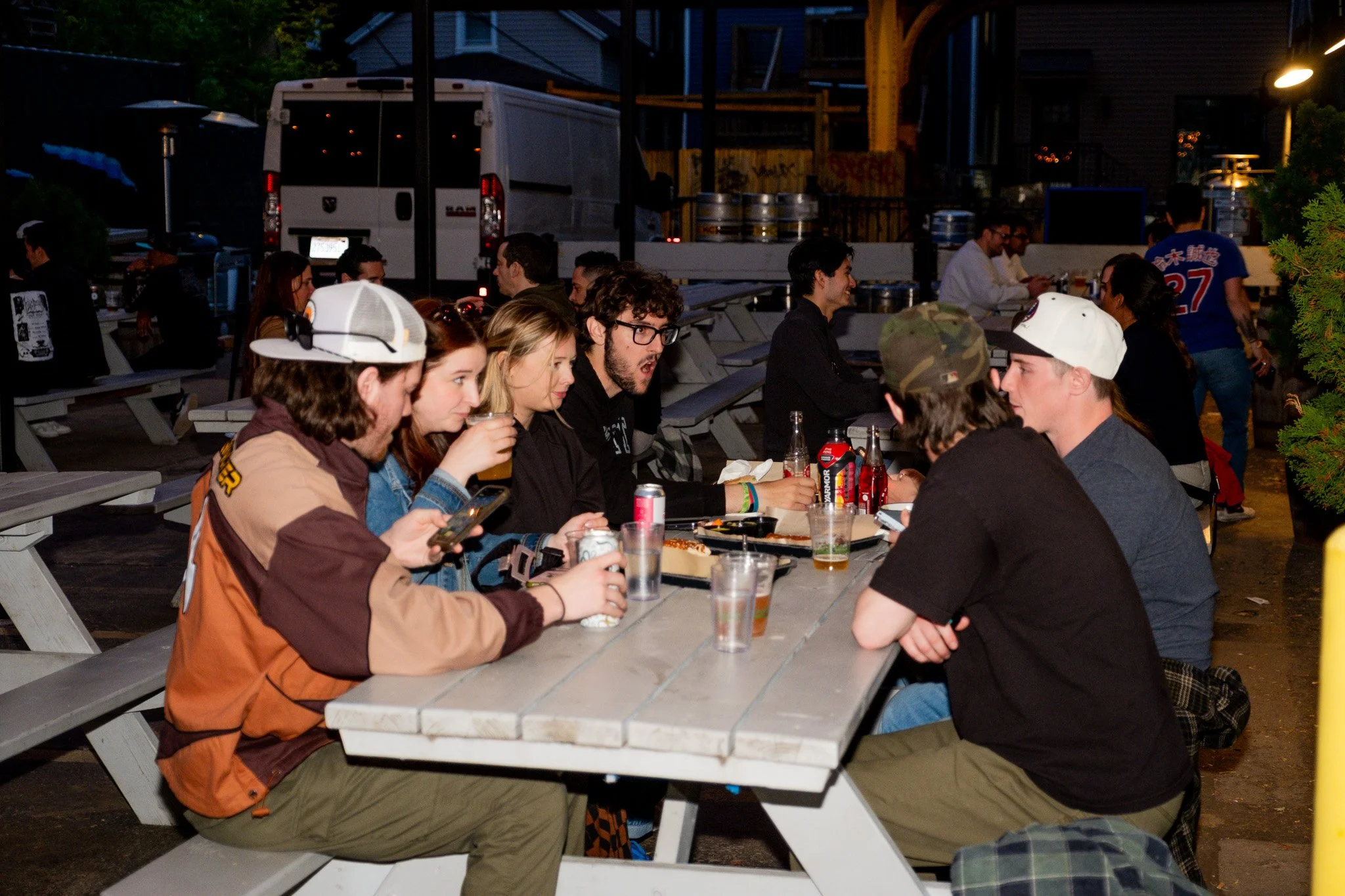 A group of young people sitting at an outdoor picnic table enjoying drinks and food, some looking at their phones, with a city street and parking area in the background at night.