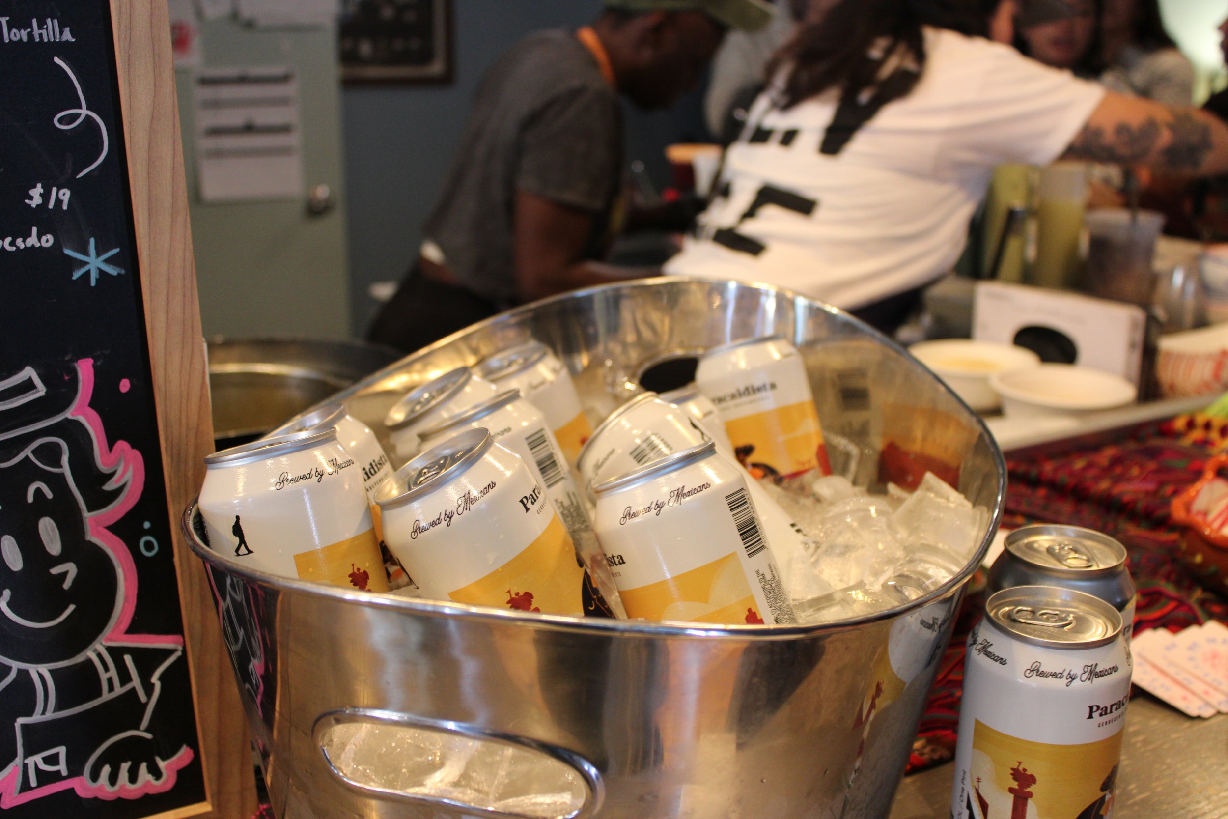 A metal bucket filled with ice and cans of beer, with some cans outside the bucket, at a bar or restaurant counter.