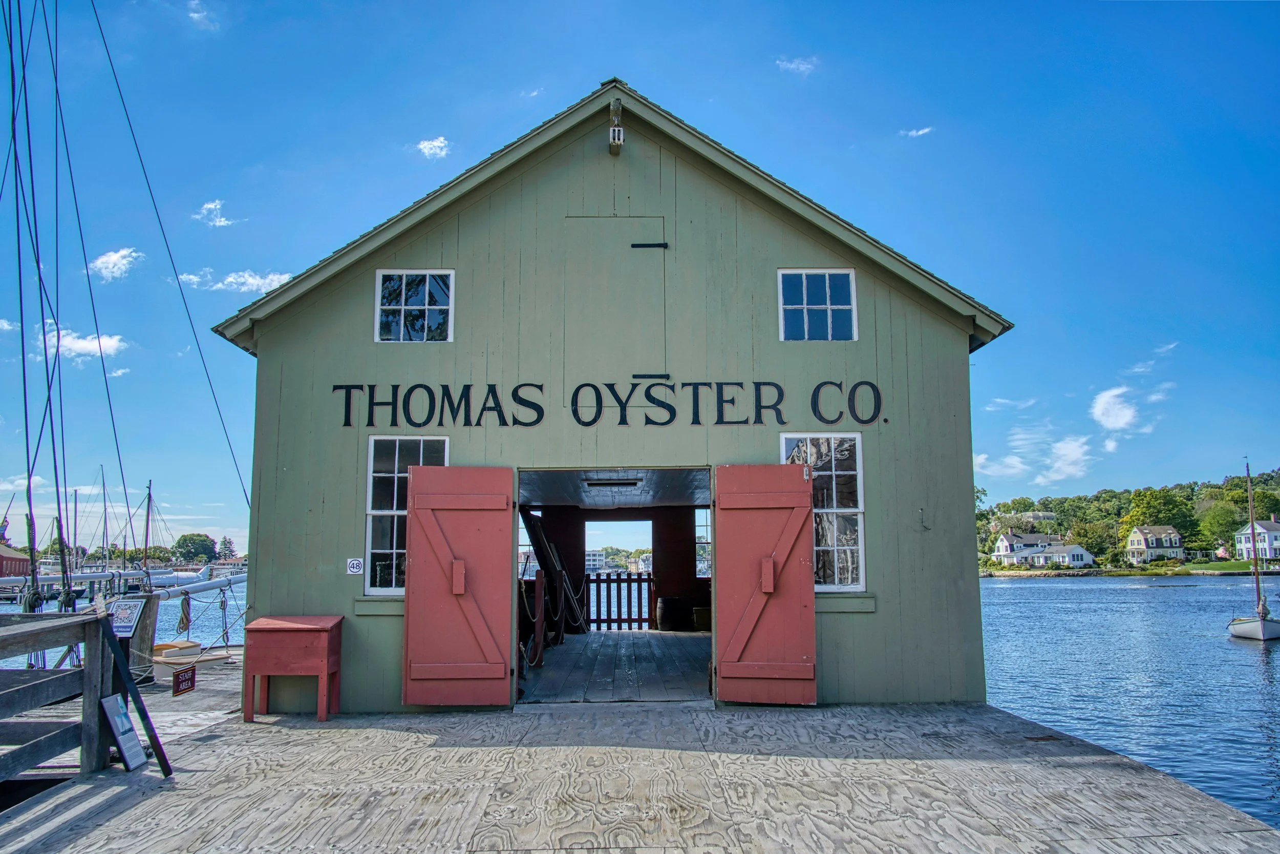 Green wooden building on a dock by the water with the sign 'Thomas Oyster Co.' above its open entrance. The building has two windows on each side and pink shutters. There are boats and houses visible across the water under a blue sky.