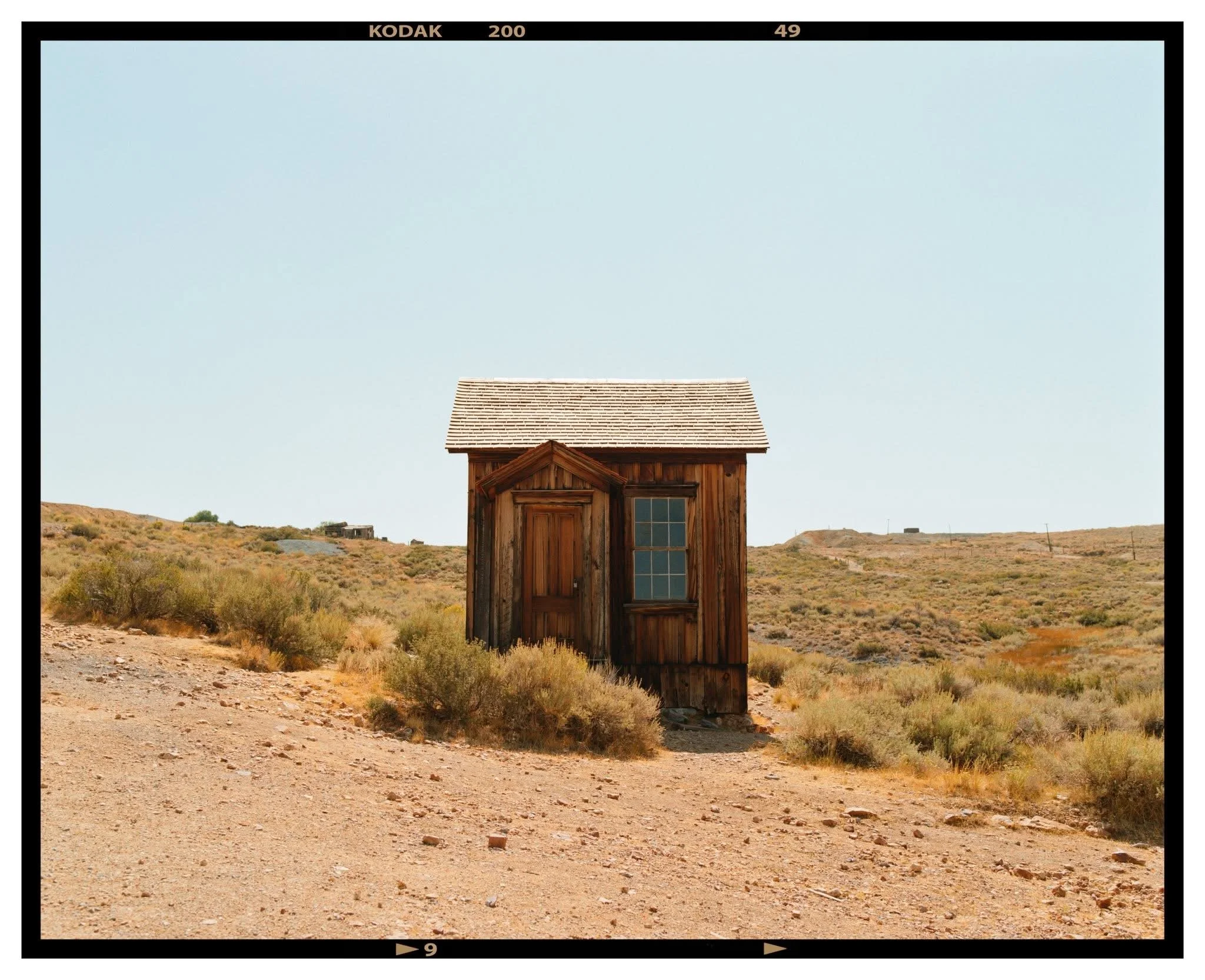 A small, rustic wooden shed standing alone in a dry, arid landscape with sparse bushes and a clear sky.