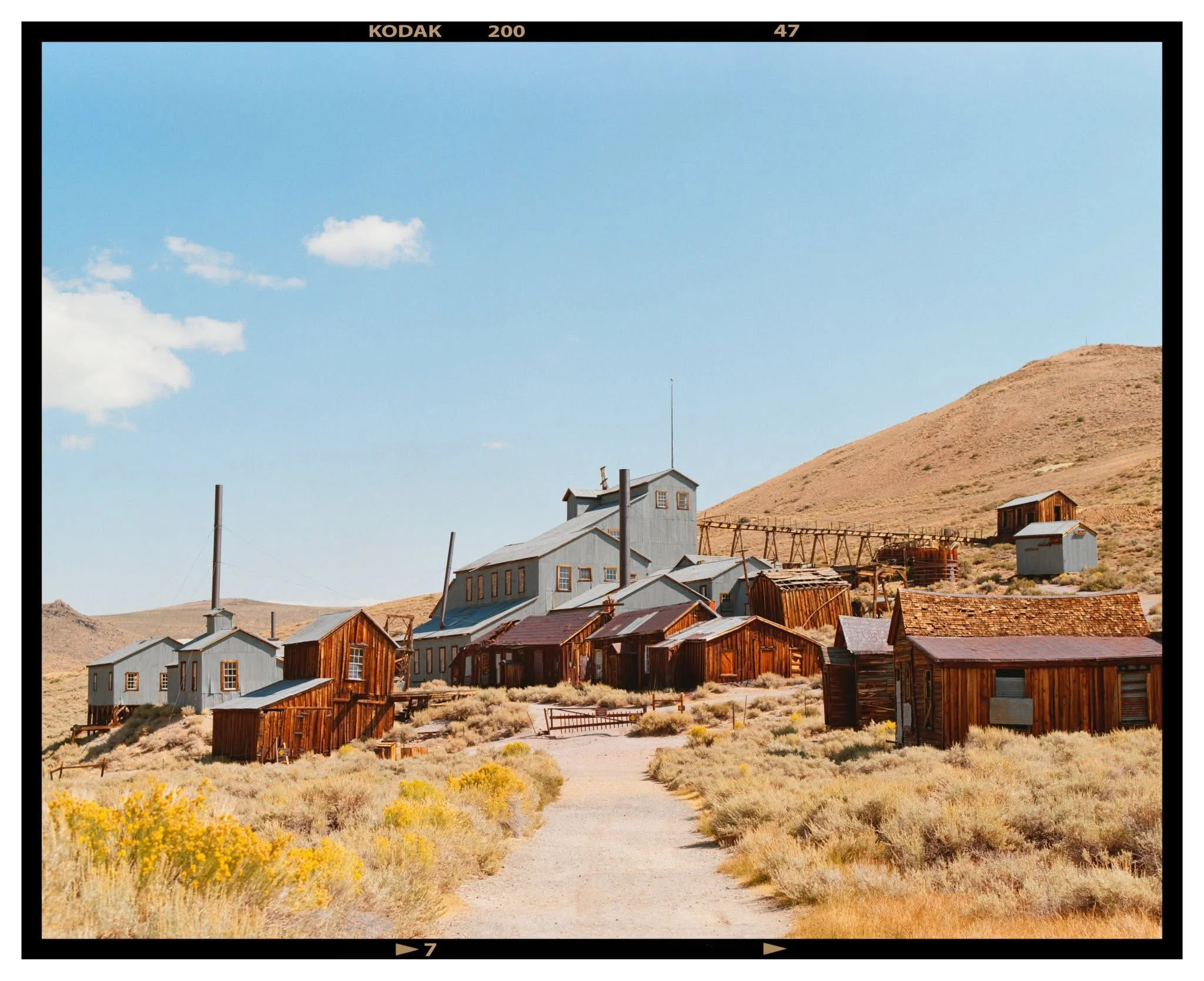 An old, abandoned mining town with wooden buildings and a large gray building on a hillside in a desert landscape, with a clear blue sky.