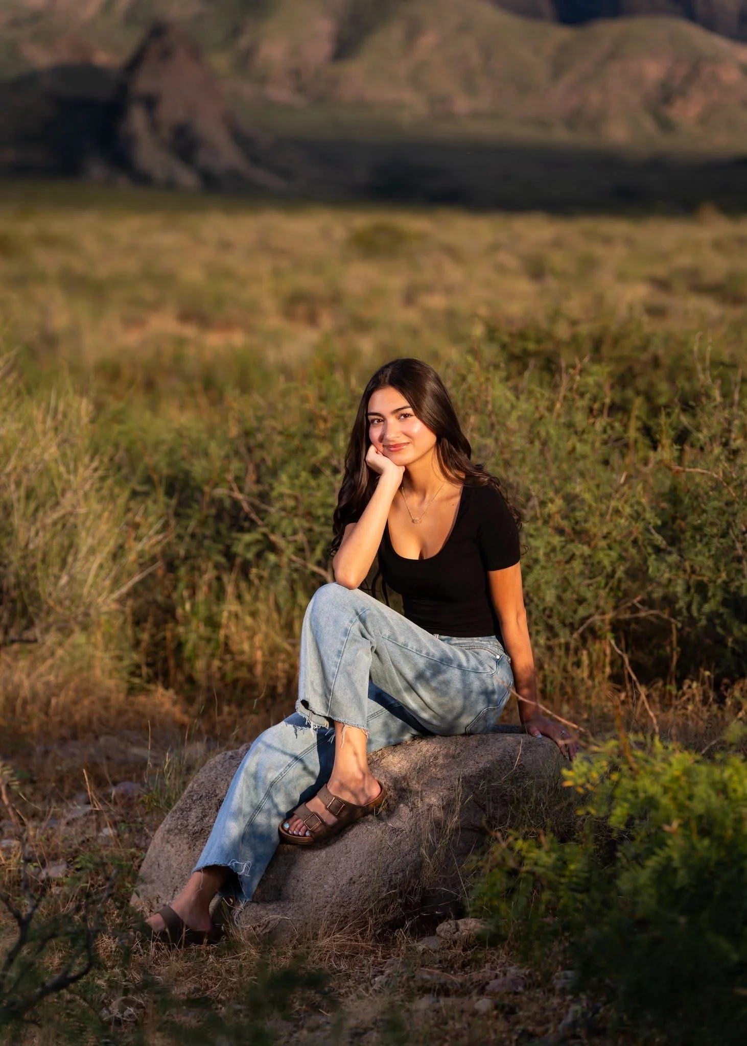 A young woman with long dark hair sitting on a large rock in a natural outdoor setting with greenery and mountains in the background, smiling at the camera.