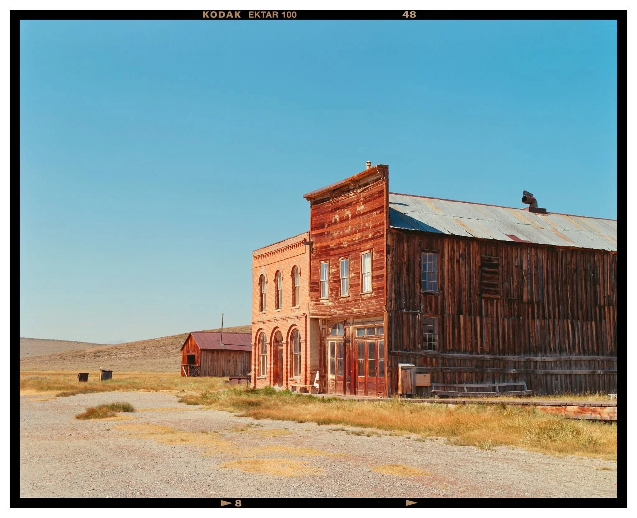An old, weathered wooden building with a brick facade on one side, situated in a rural, open landscape with grassy fields and rolling hills under a clear blue sky.