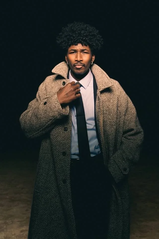 A young man with curly hair wearing a tan herringbone coat over a light blue shirt and dark tie, standing outdoors at night, posing confidently with one hand on his chest.
