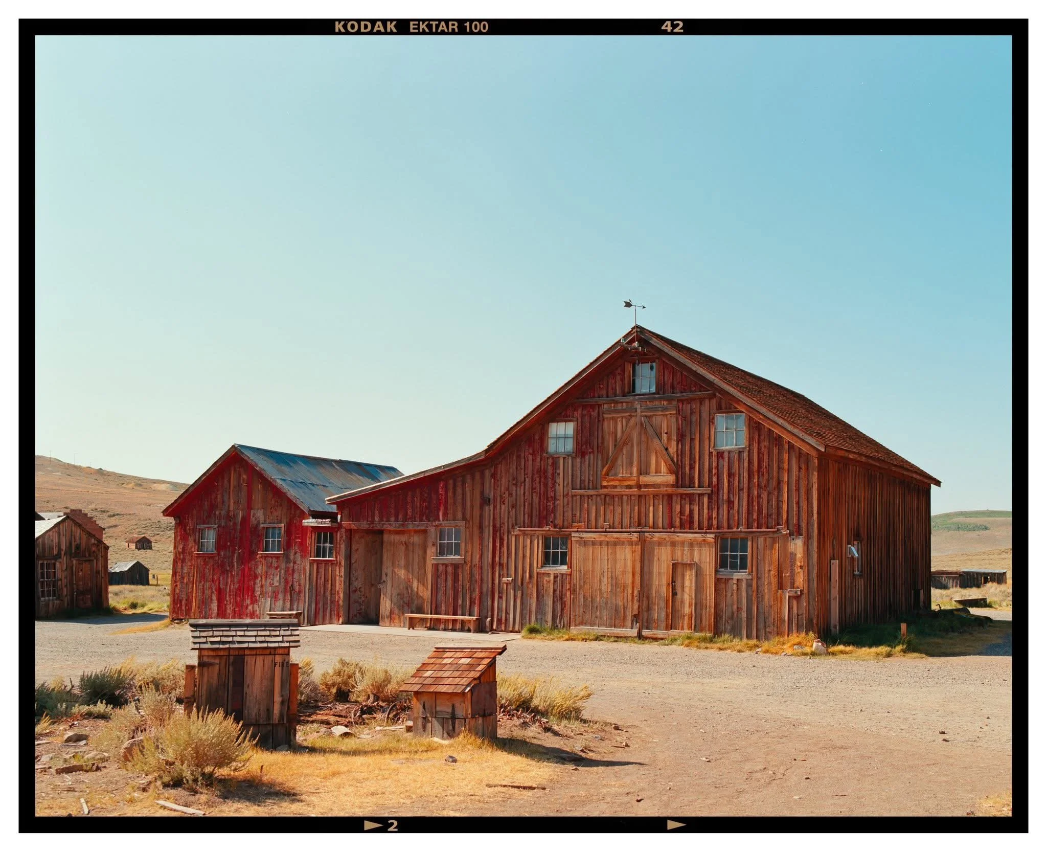 A large red wooden barn with a smaller attached shed, situated in a rural area with dry grass and hills in the background, under a clear blue sky.
