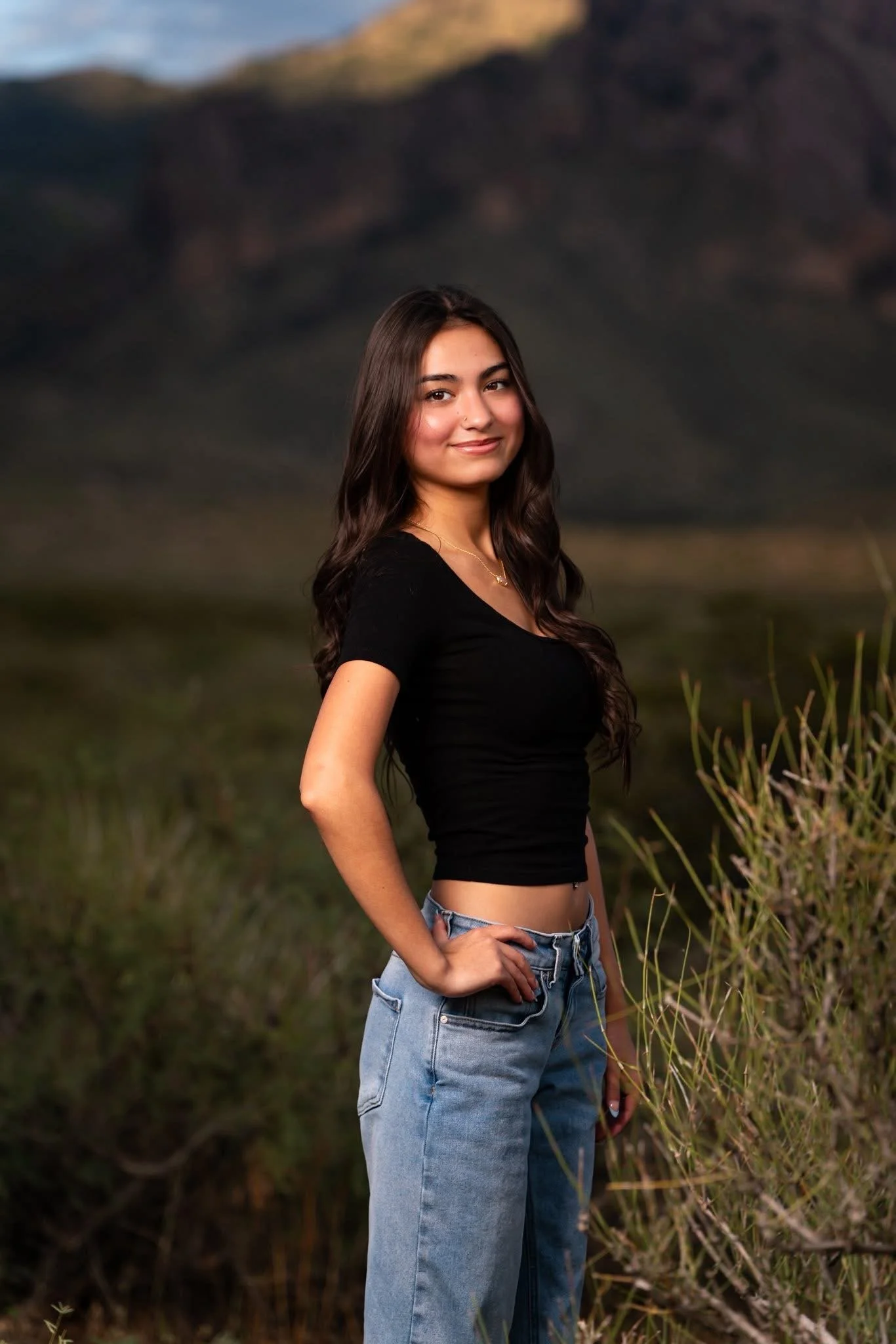 A young woman with long dark hair wearing a black crop top and light blue jeans standing outdoors with a mountainous landscape in the background.