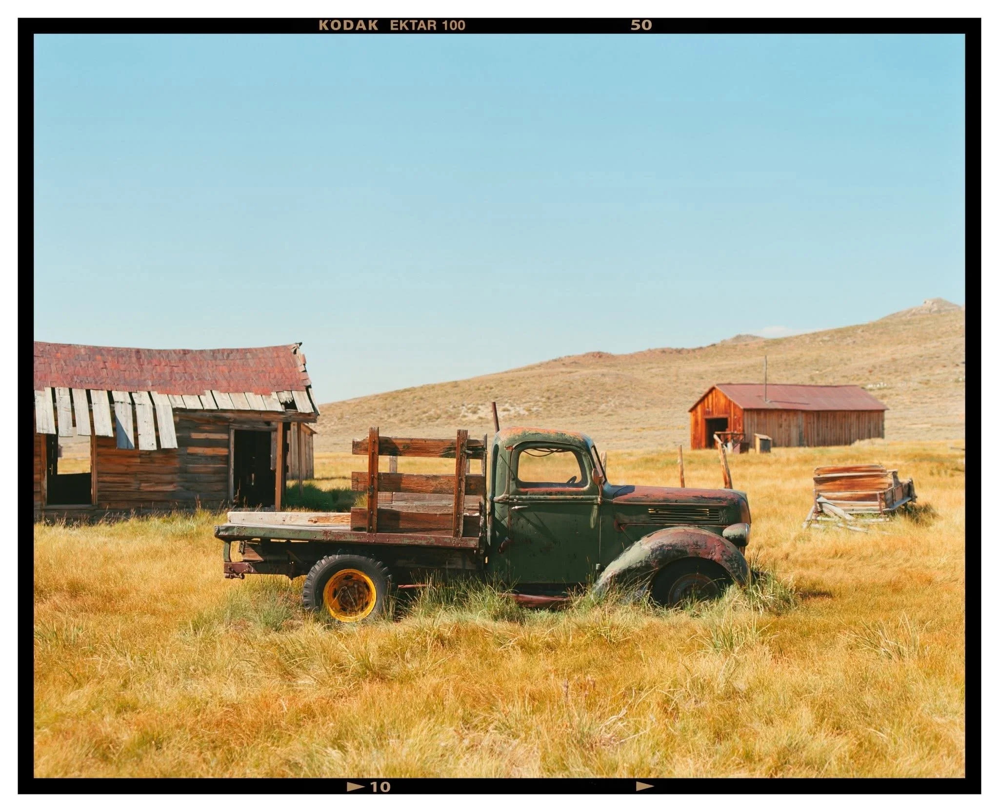 An abandoned vintage green pickup truck in a grassy field, with dilapidated wooden farm buildings and a landscape of rolling hills under a clear blue sky in the background.