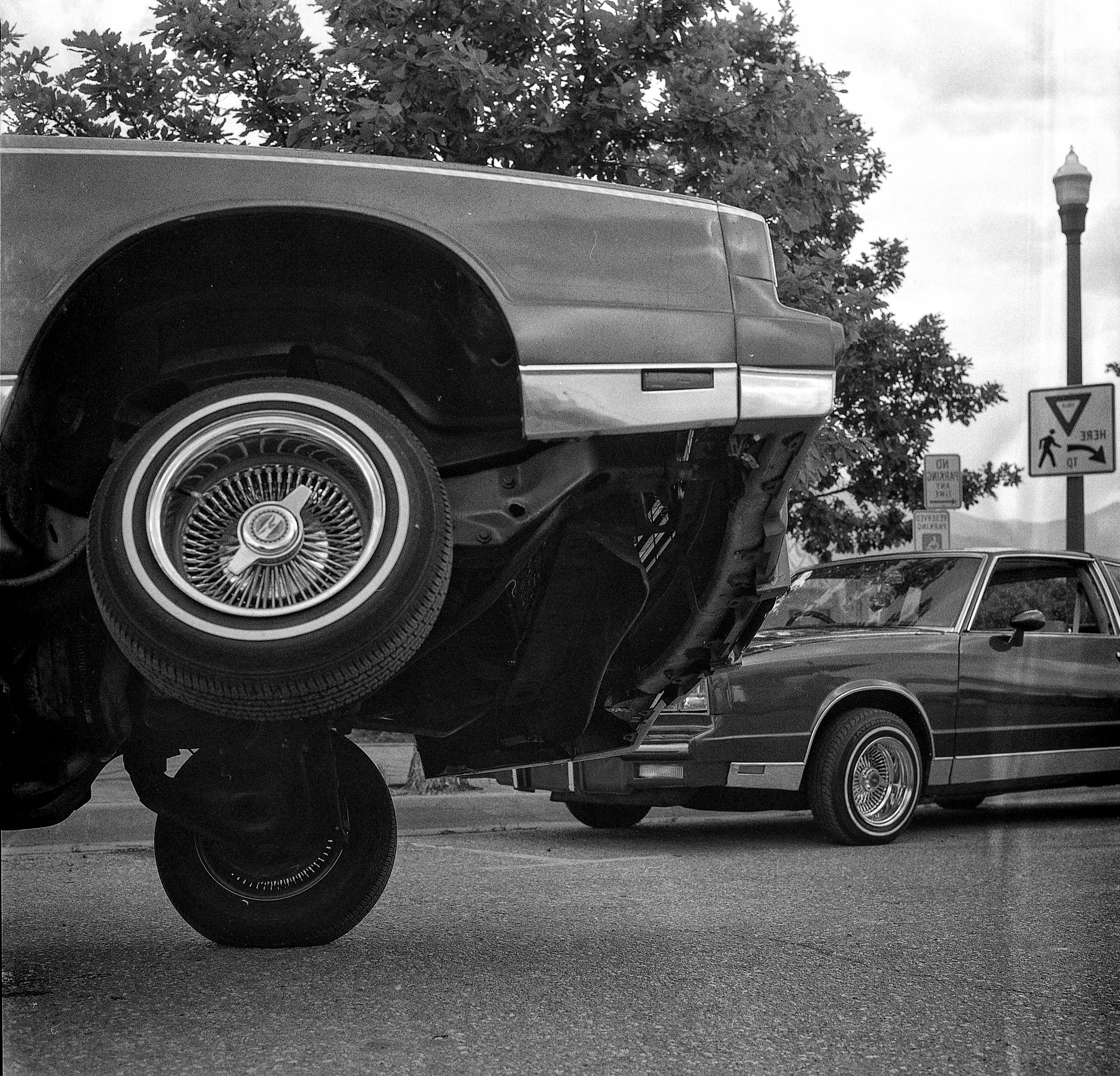 Two cars parked on the street, with one car's front end elevated on a jack stand. The sky is cloudy, and trees and street signs are visible in the background.