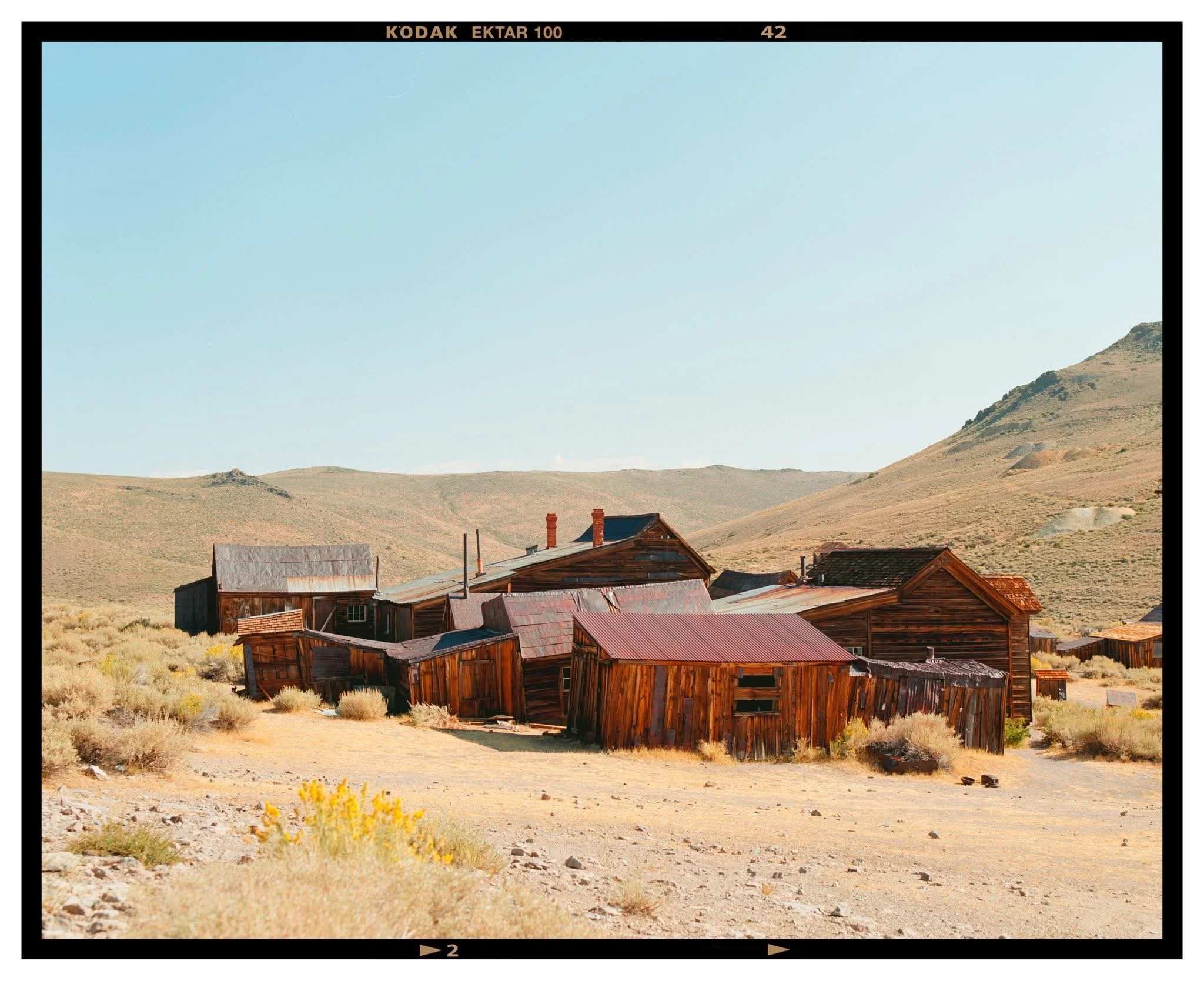 Old, weathered wooden buildings in a desert landscape with rolling hills and clear blue sky.