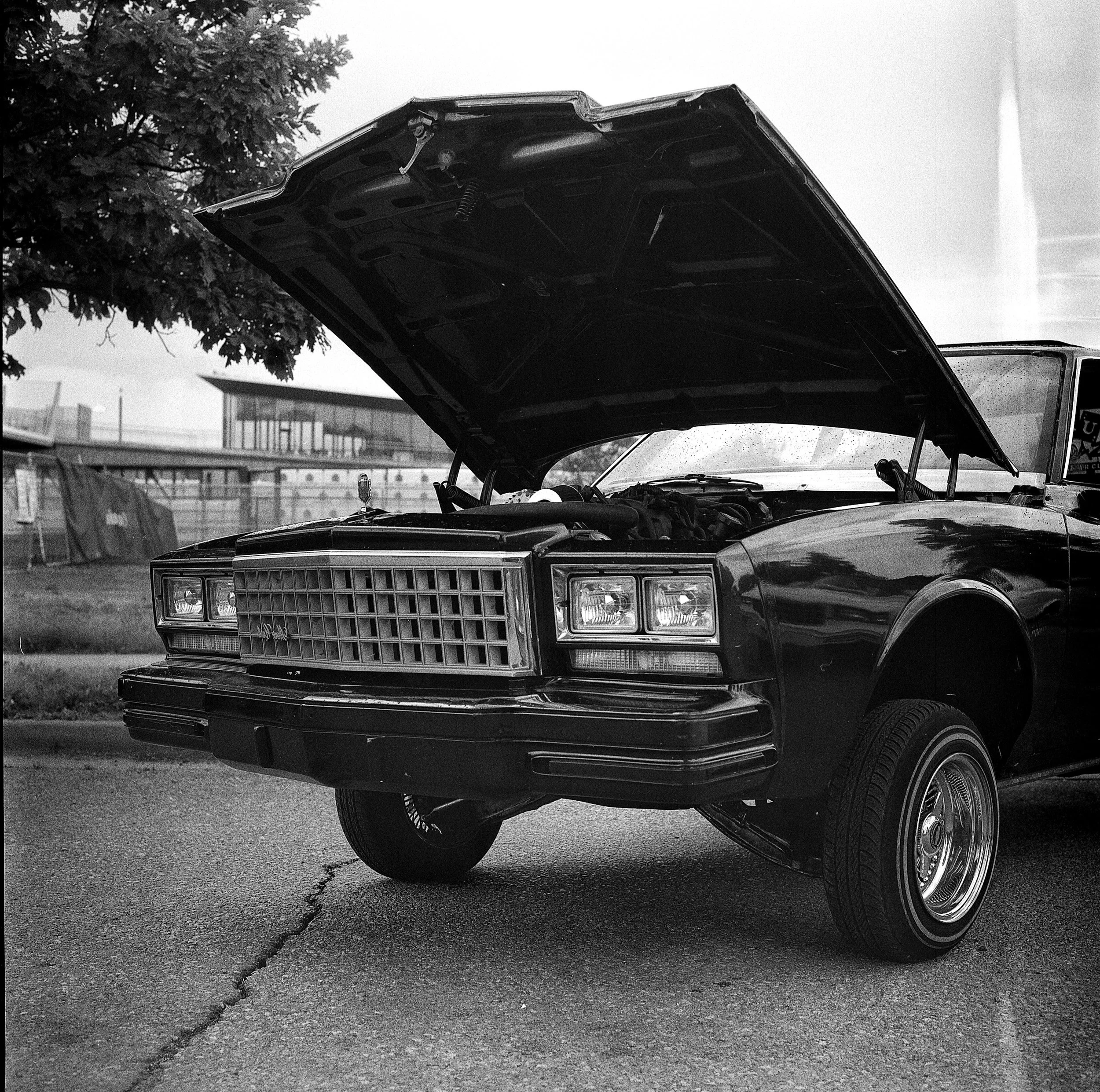 Black vintage car with open hood, parked on street, showing engine, with a tree and building in background.