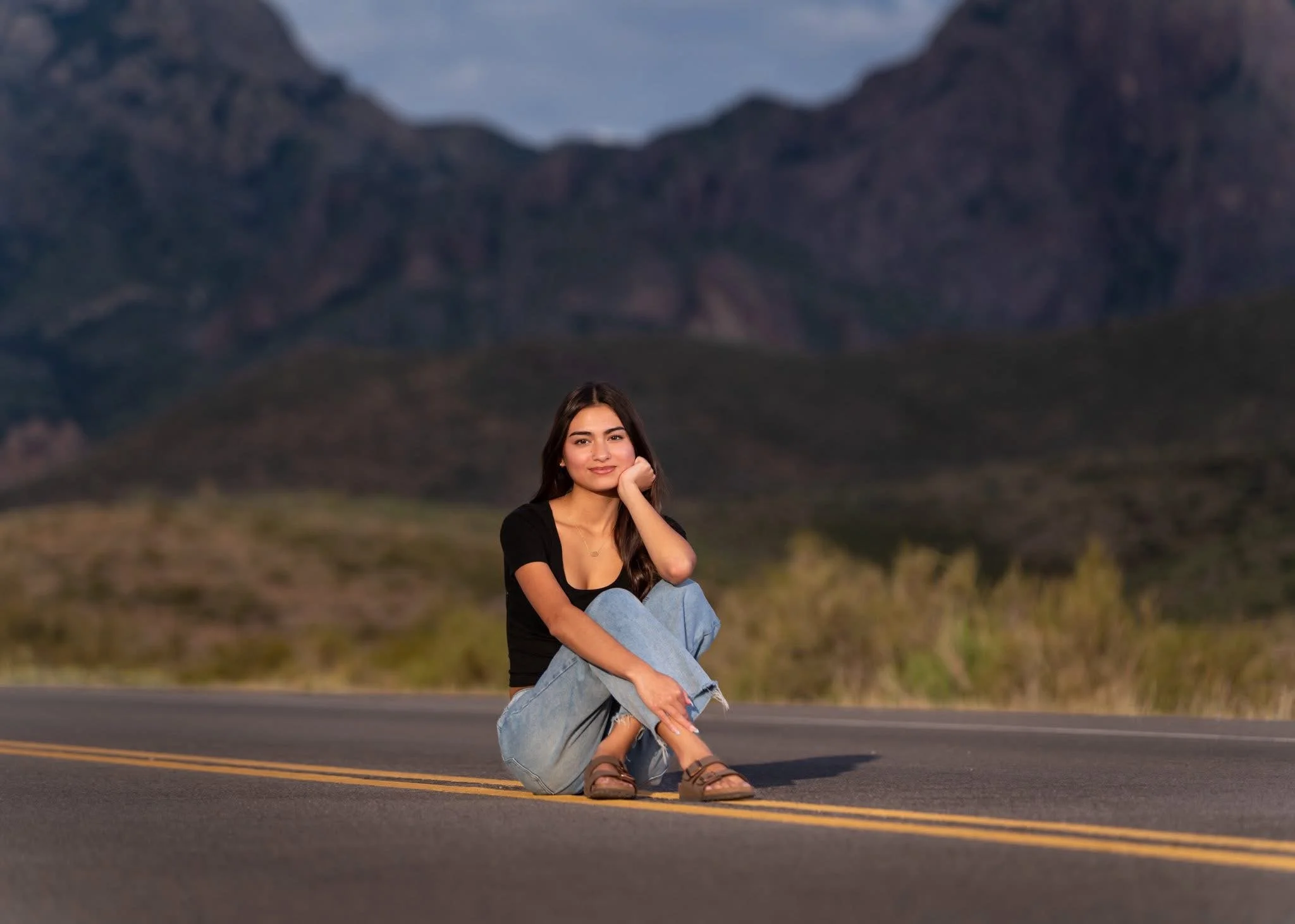 A young woman sitting on an empty road in a rural area with mountains in the background, wearing a black t-shirt, jeans, and sandals, resting her chin on her hand and smiling.