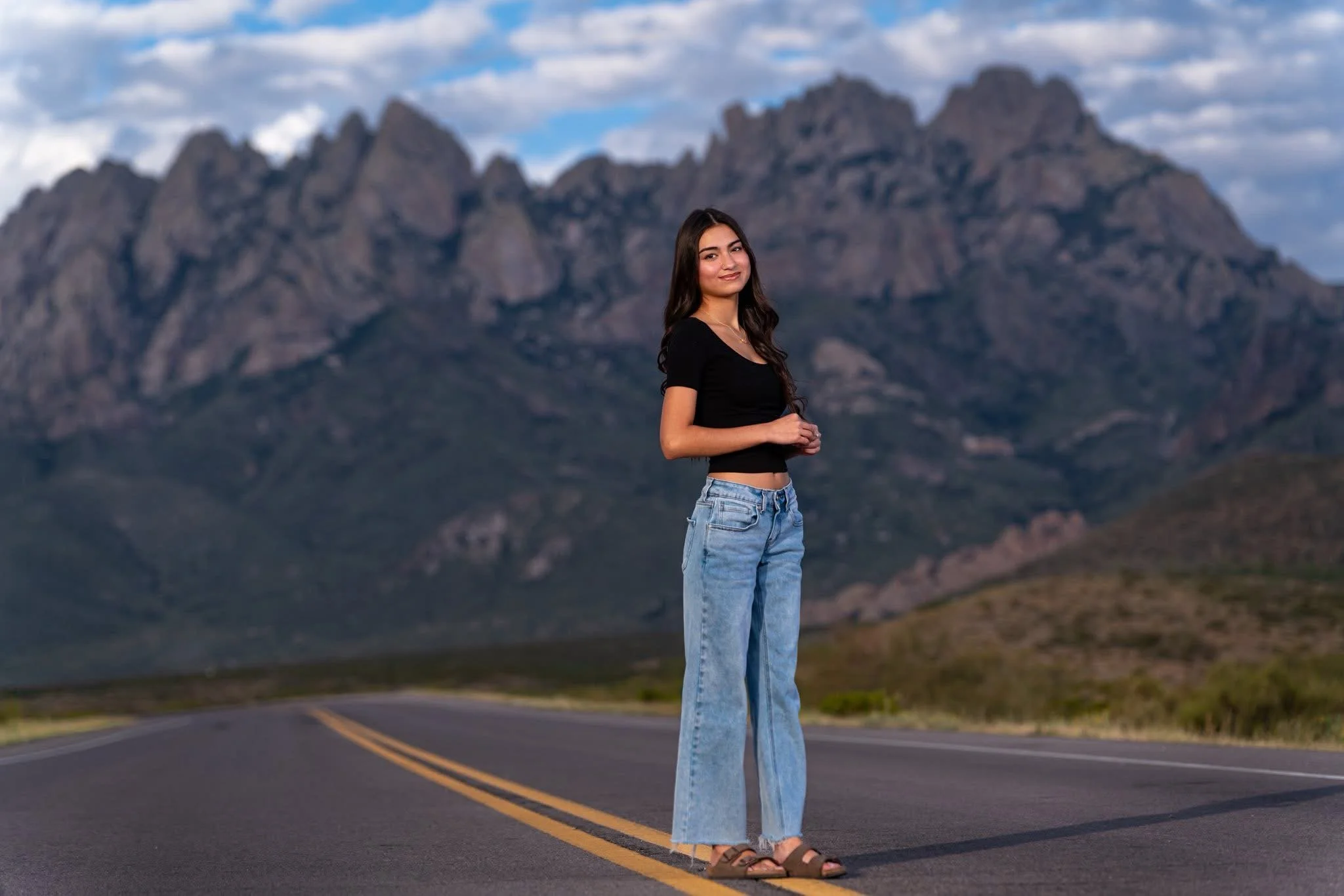 A young woman standing on an empty road with mountains in the background, wearing a black crop top, jeans, and sandals, looking at the camera.