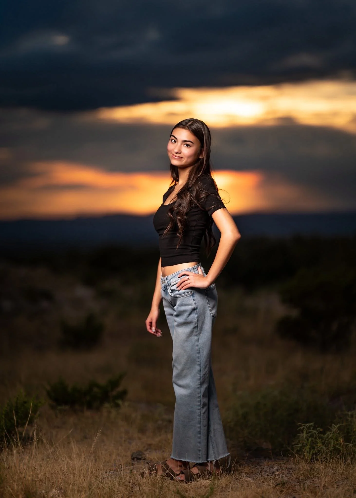 A young woman stands outdoors during sunset, wearing a black crop top and light-wash jeans, with her hand on her hip and smiling at the camera.
