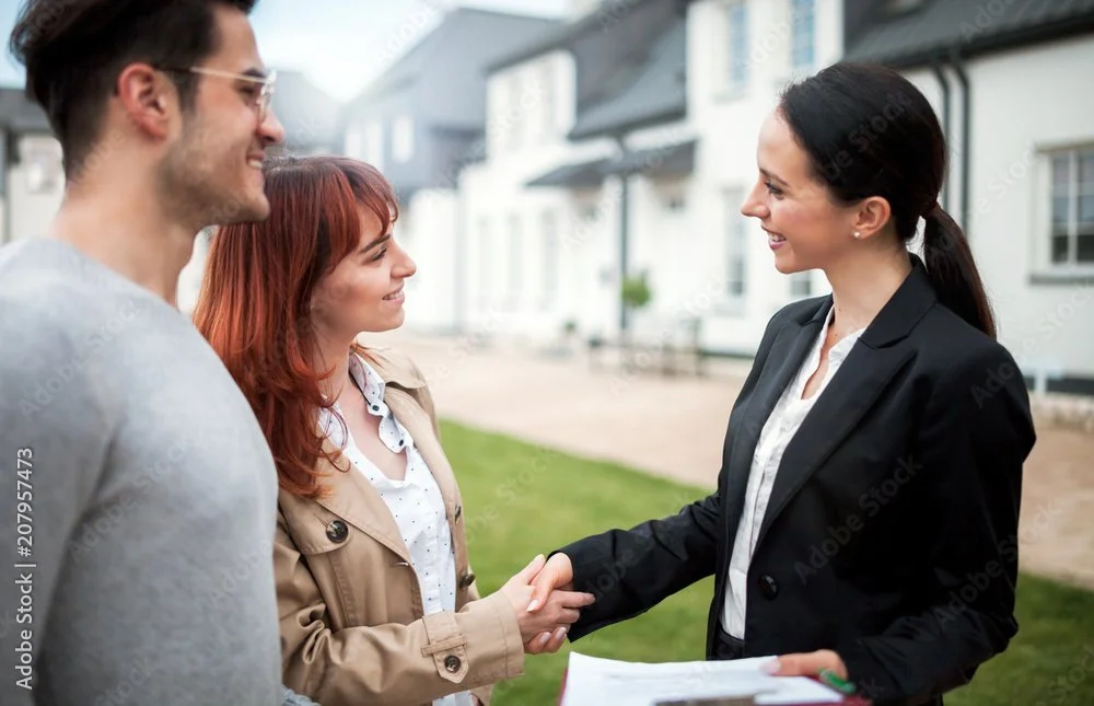 A Real Estate Professional in a navy blazer shaking hands with Buyers in a beige trench coat outdoors, smiling, with a man in a gray sweater standing nearby looking for their new home.