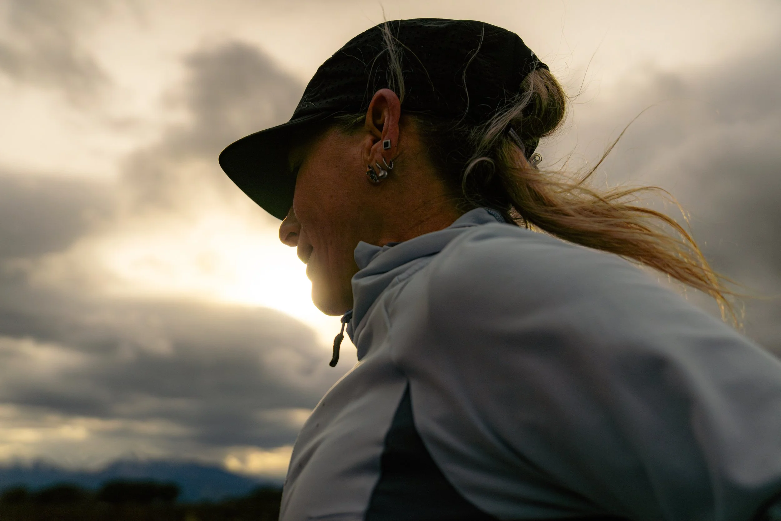 Profil d'une femme portant une casquette noire, des boucles d'oreilles en plusieurs parties, un sweat blanc, face au couché de soleil avec un ciel nuageux.