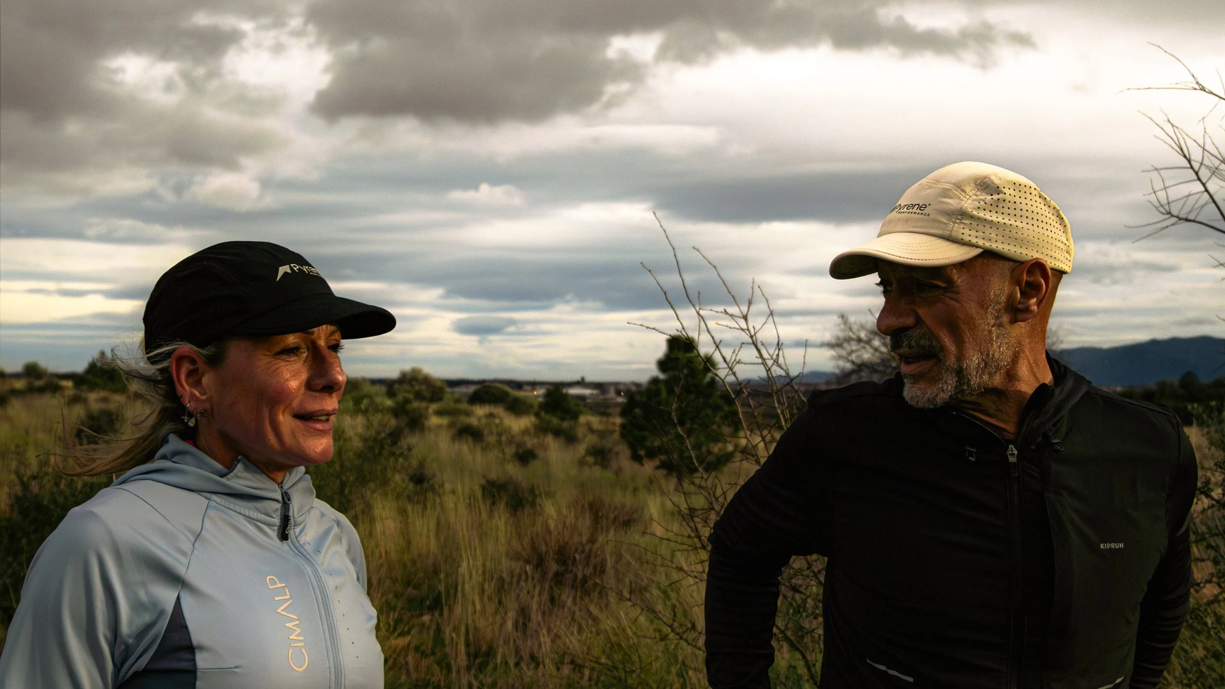 Deux personnes, un homme et une femme, parlent dans une nature herbeuse sous un ciel nuageux, vêtues de vêtements de sport, avec des montagnes en arrière-plan.