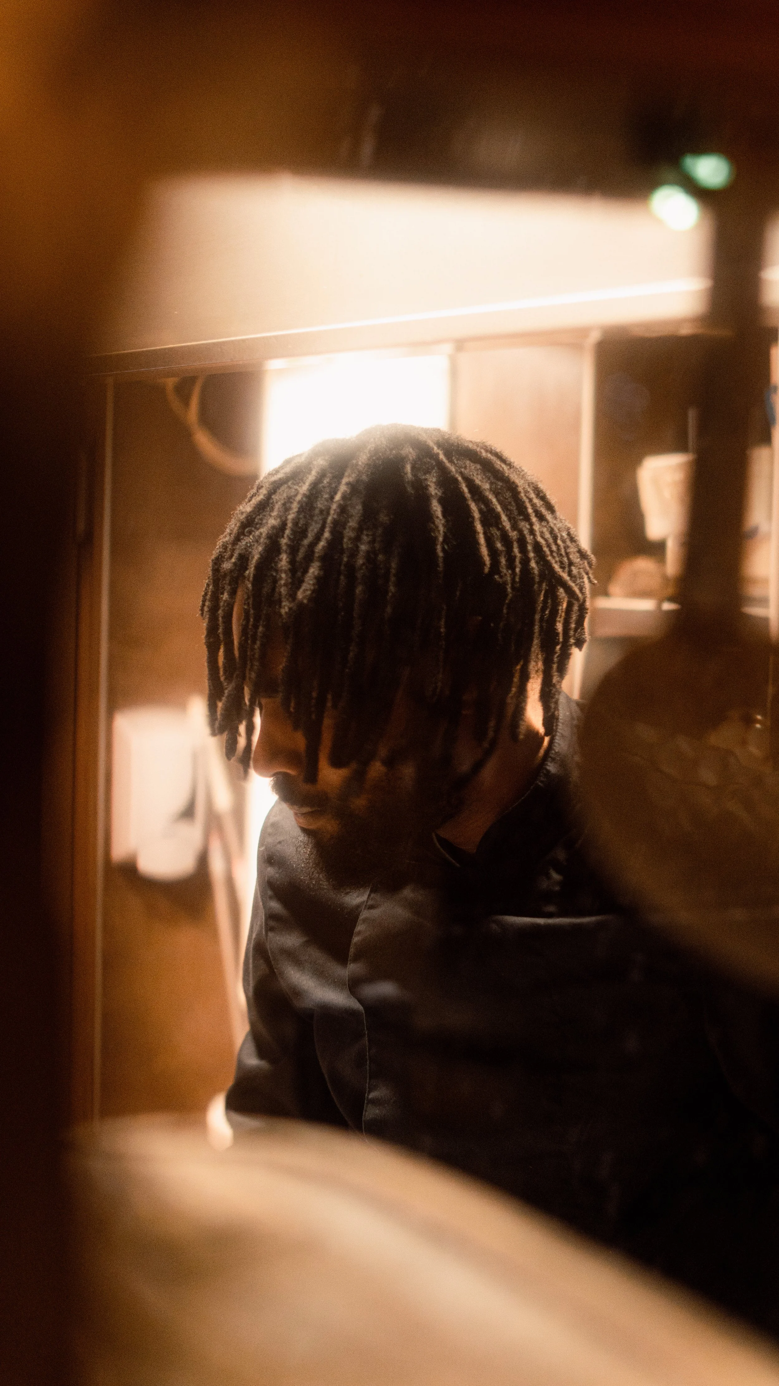 Jeune cuisinier, homme aux cheveux dreadlocks, portant une veste noire, regardant vers le bas, dans une cuisine intérieur tamisé.