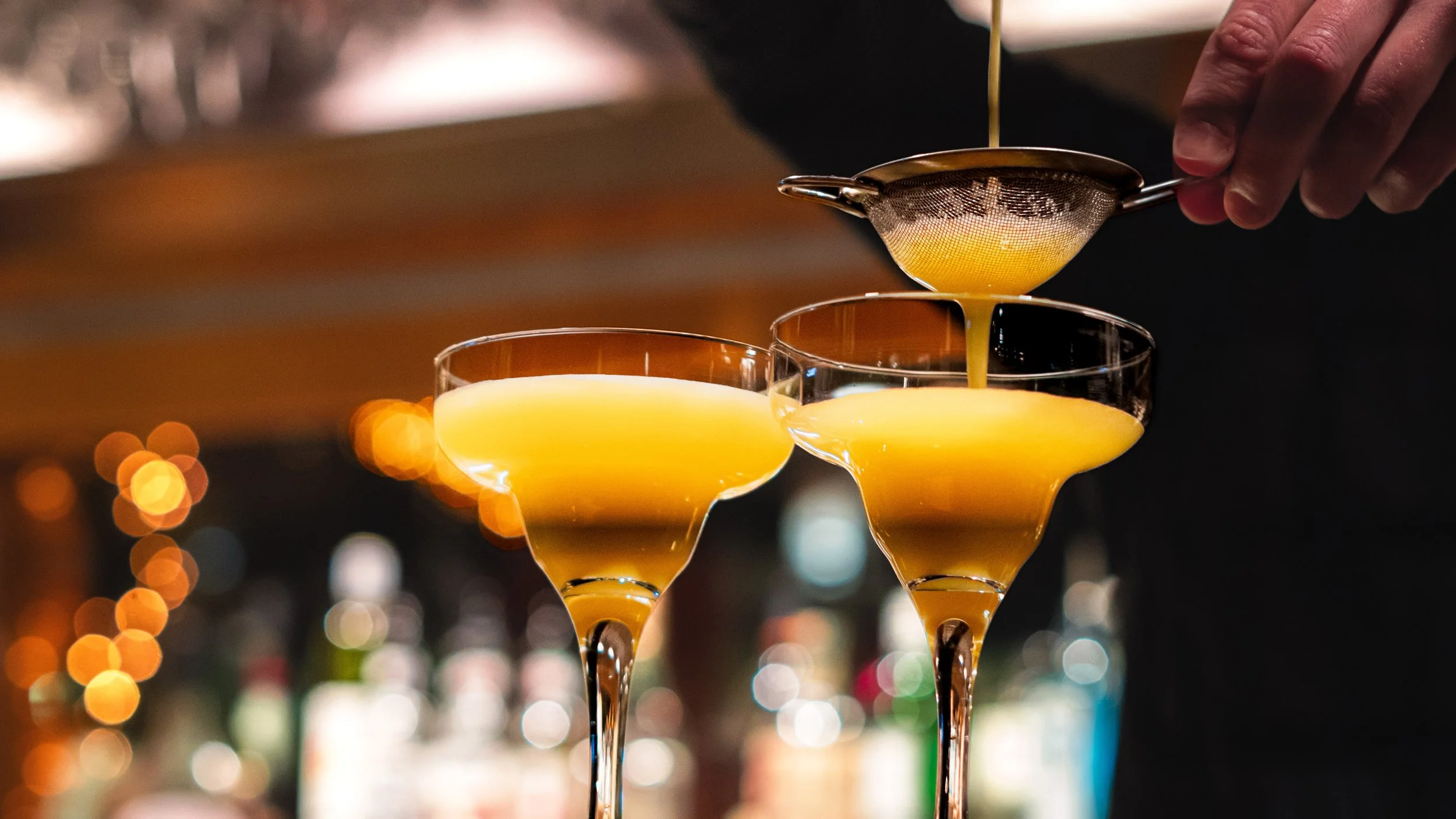 Un barman verse un liquide jaune dans des verres à cocktail, de formant coupe, avec un fond flou de lumières chaudes.