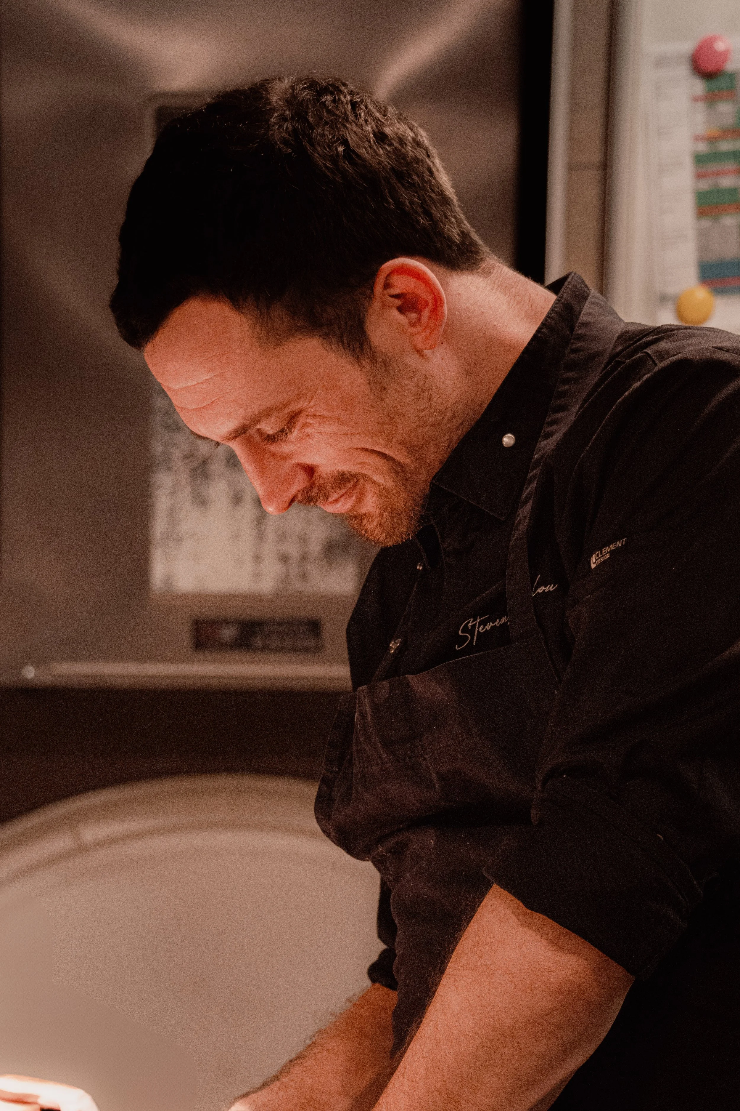 Un homme souriant, portant un tablier noir, en train de cuisiner dans une cuisine.