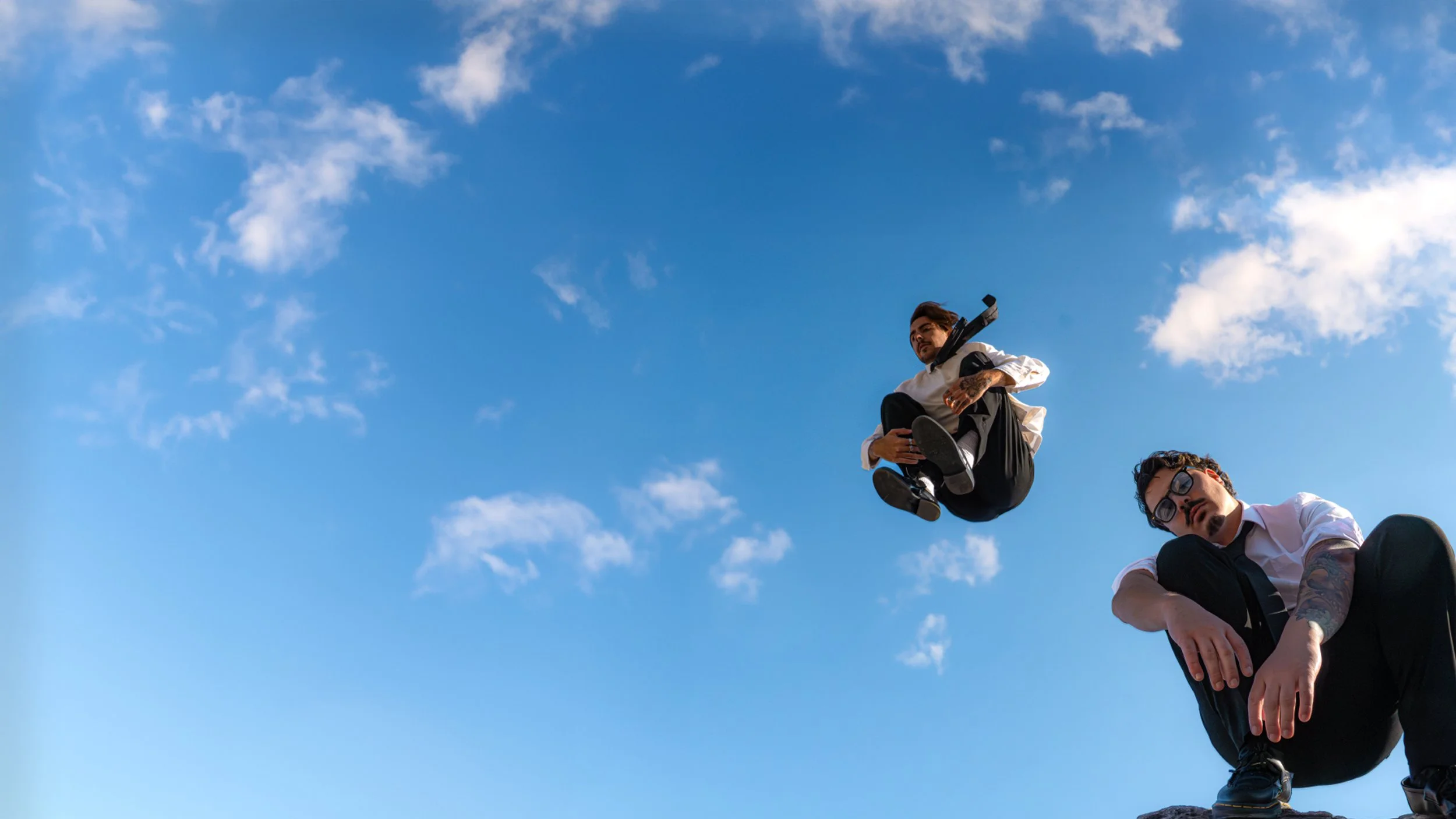 Deux hommes en costume noir et blanc, avec des tatouages, assis sur une roche contre un ciel bleu avec quelques nuages.
