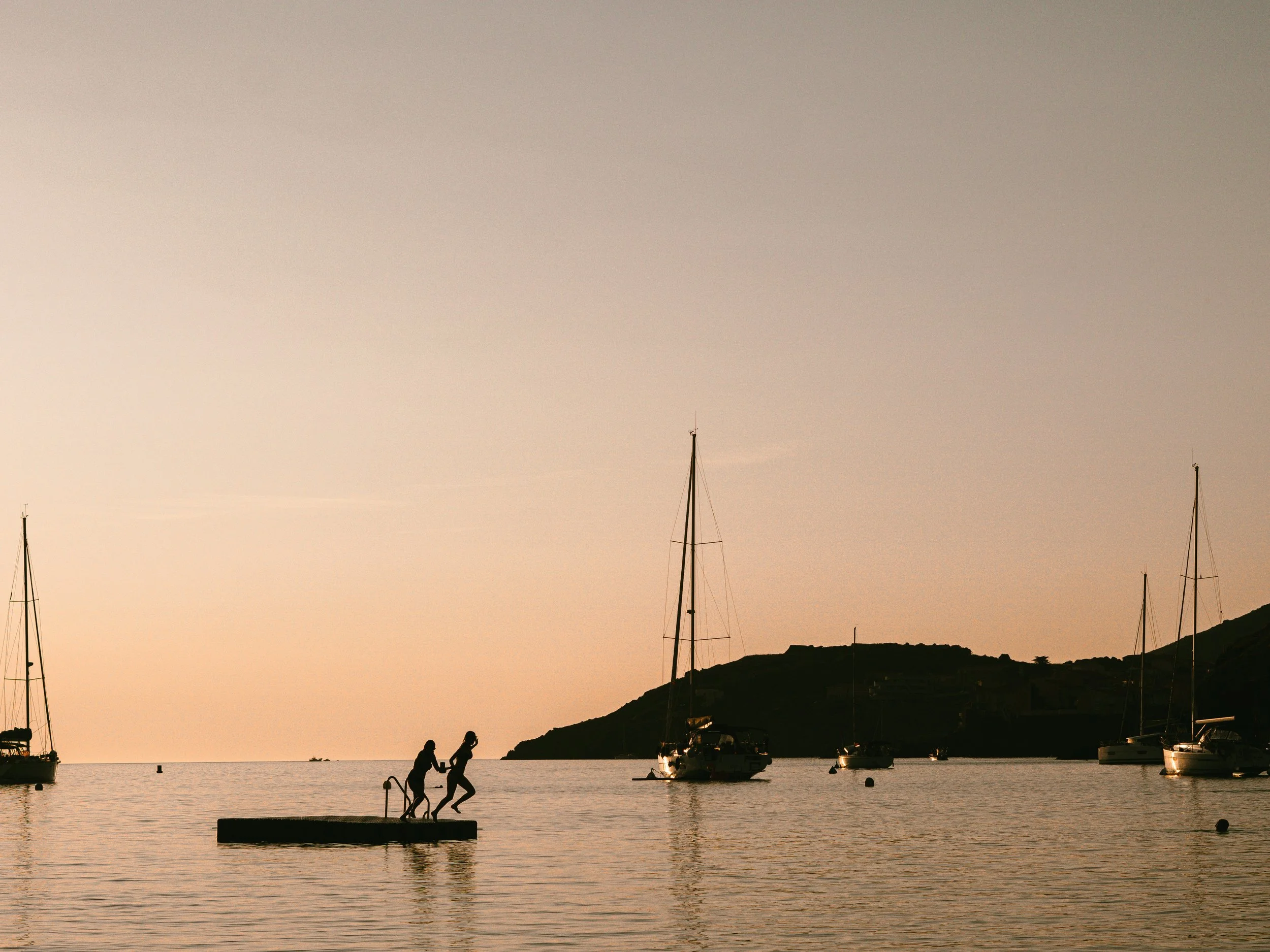 Couchers de soleil sur un port avec des voiliers, deux personnes sautant dans l'eau sur une plateforme.