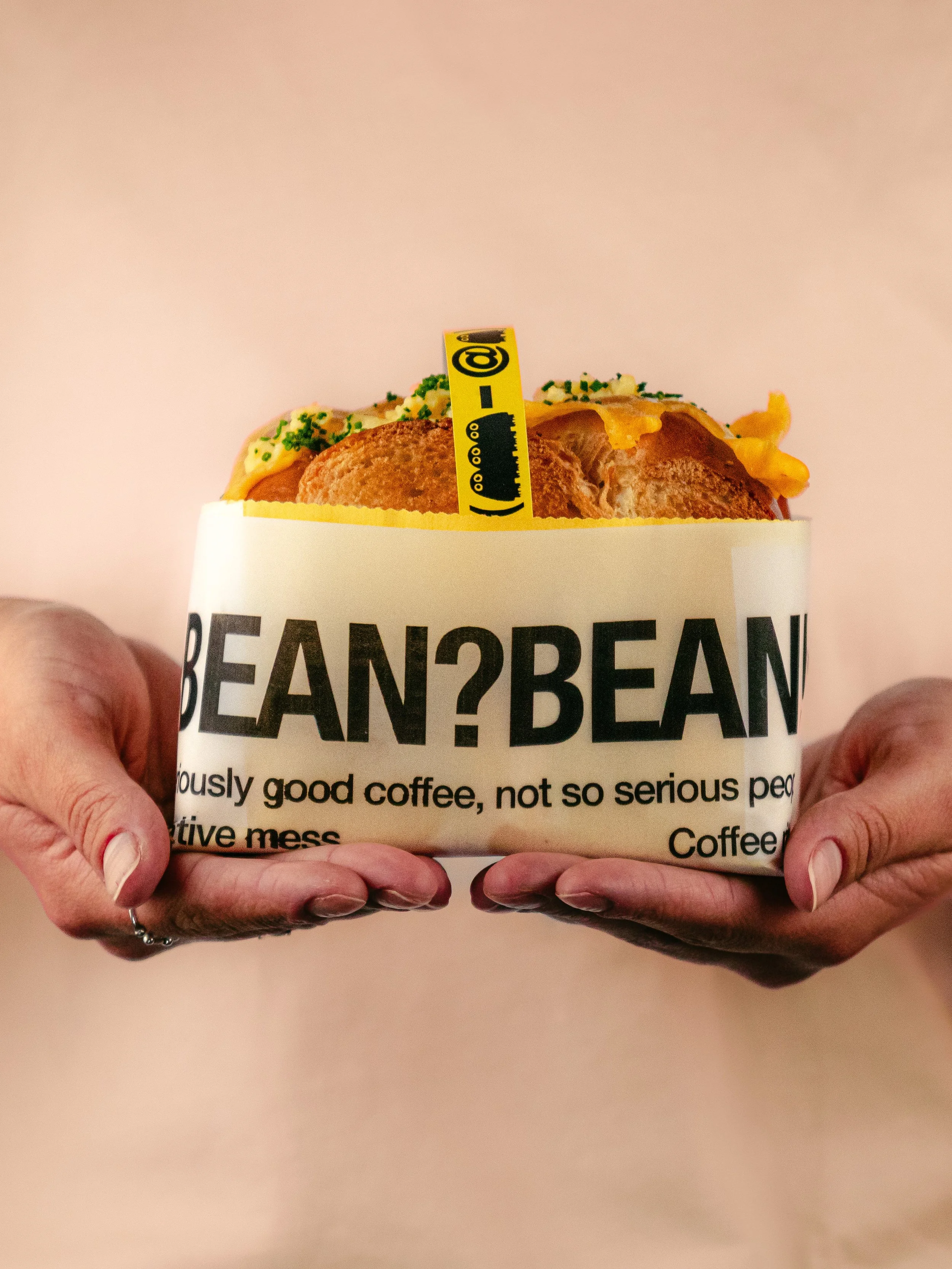 Un sandwich chaud avec un haut parleur jaune avec symbole @, dans une boîte en papier blanc avec le texte 'BEAN?BEAN' et des mots en anglais.