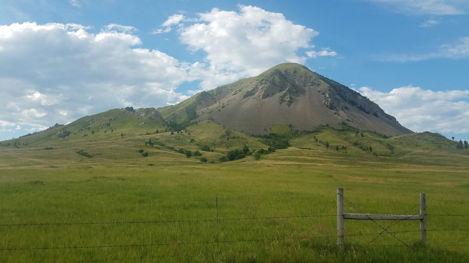 A scenic landscape featuring a large, grassy plain with a mountain in the background. The sky is partly cloudy with patches of blue. There is a simple wooden fence in the foreground.