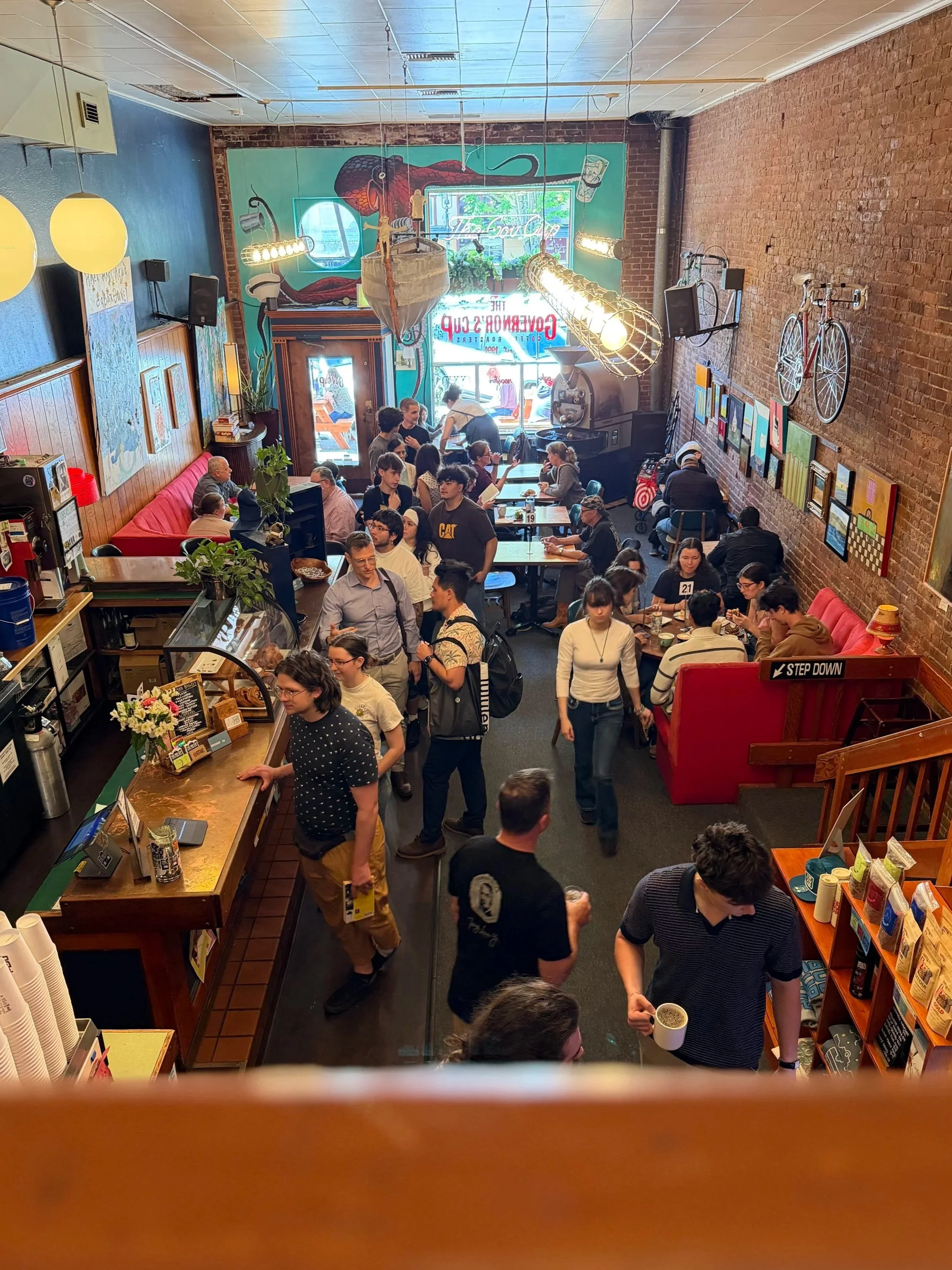 Inside a busy cafe filled with people sitting at tables and standing near the counter. Decor includes a large mural of a snake on the back wall, artwork on the side walls, a bicycle mounted on the brick wall, and various hanging light fixtures. There are plants and a neon sign outside the front window.
