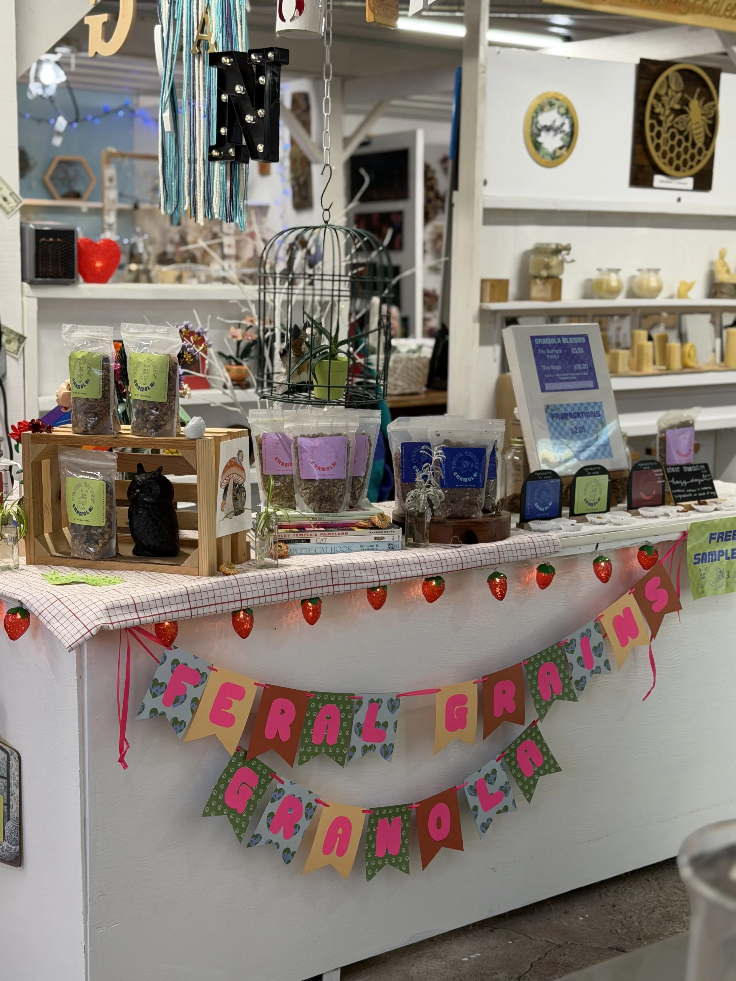 A table decorated with a Strawberry Graffiti themed banner, small potted plants, packaged products labeled 'Graffiti,' and a sign with prices. In the background, shelves with various decorative items and a monitor are visible.