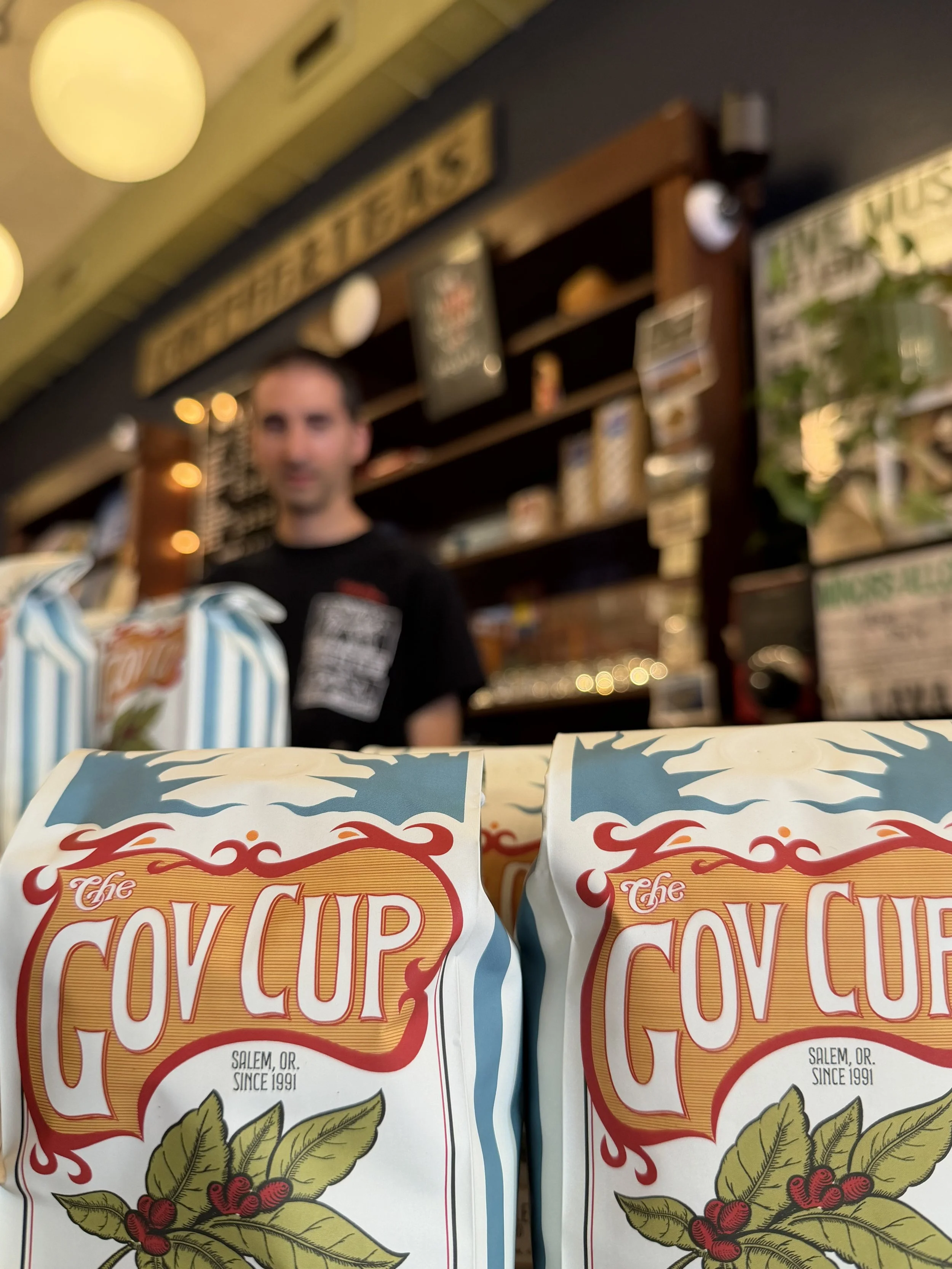 Two bags of 'The Gov Cup,' coffee beans in the foreground with a person and a store background behind them.