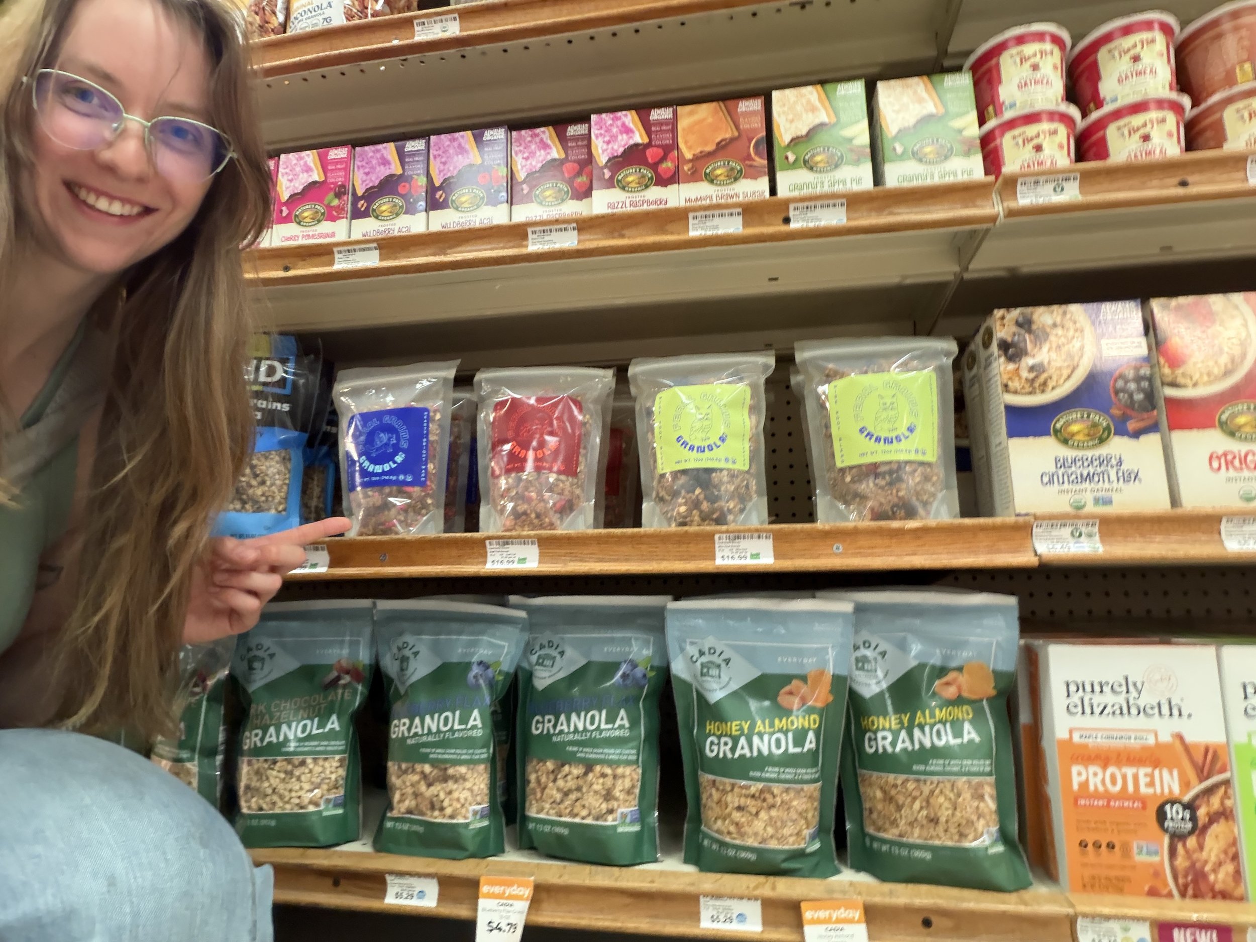 The owner of Feral Grains Granola Co. pointing at her shelf of granola at LifeSource Natural Grocers.