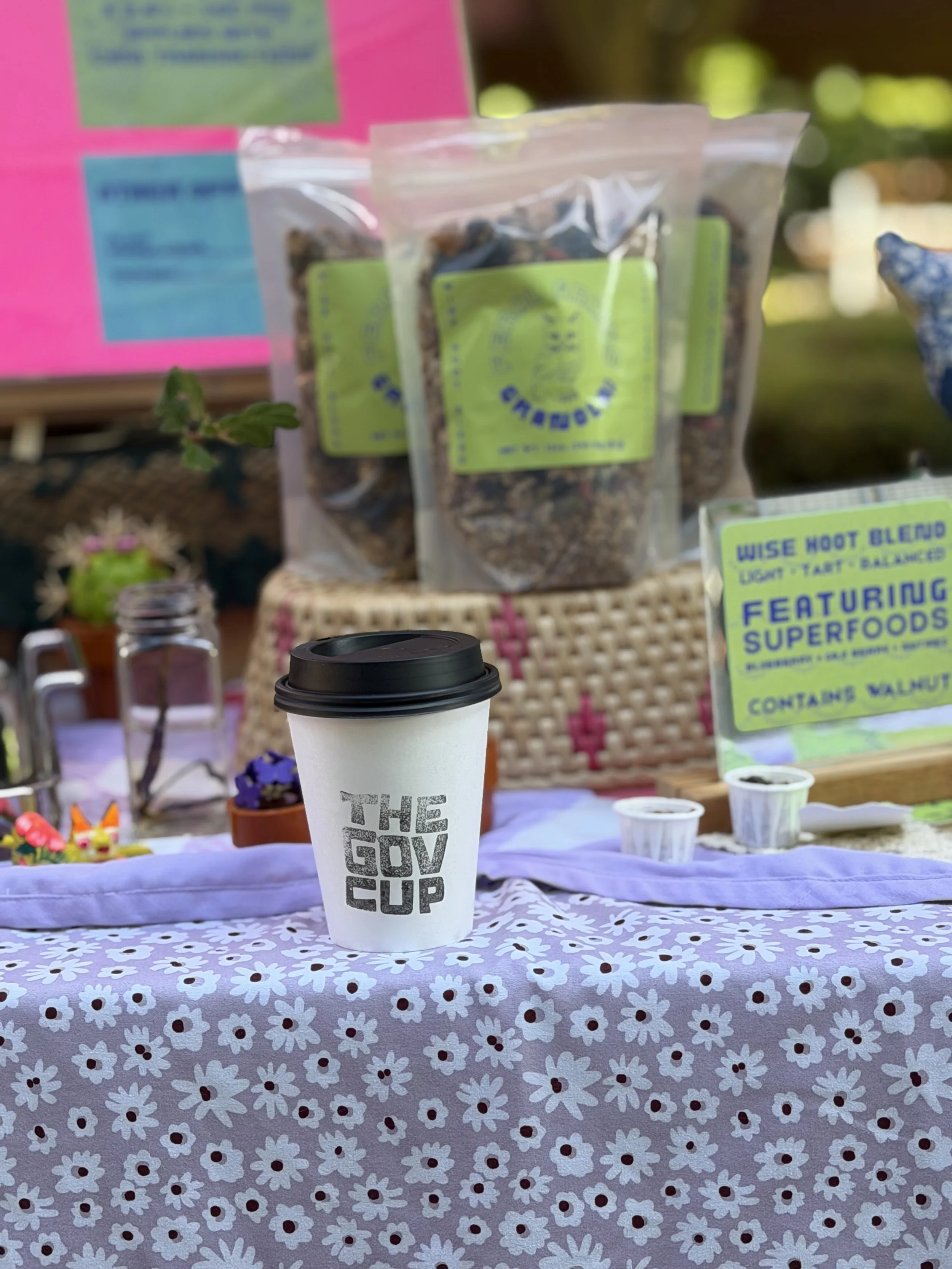 A white disposable coffee cup with black lid and the words 'THE GOV CUP' written on it, placed on a table cover with a white daisy pattern. In the background, there are various products and signs displayed on a pink backdrop, and a purple container with small items on the table.