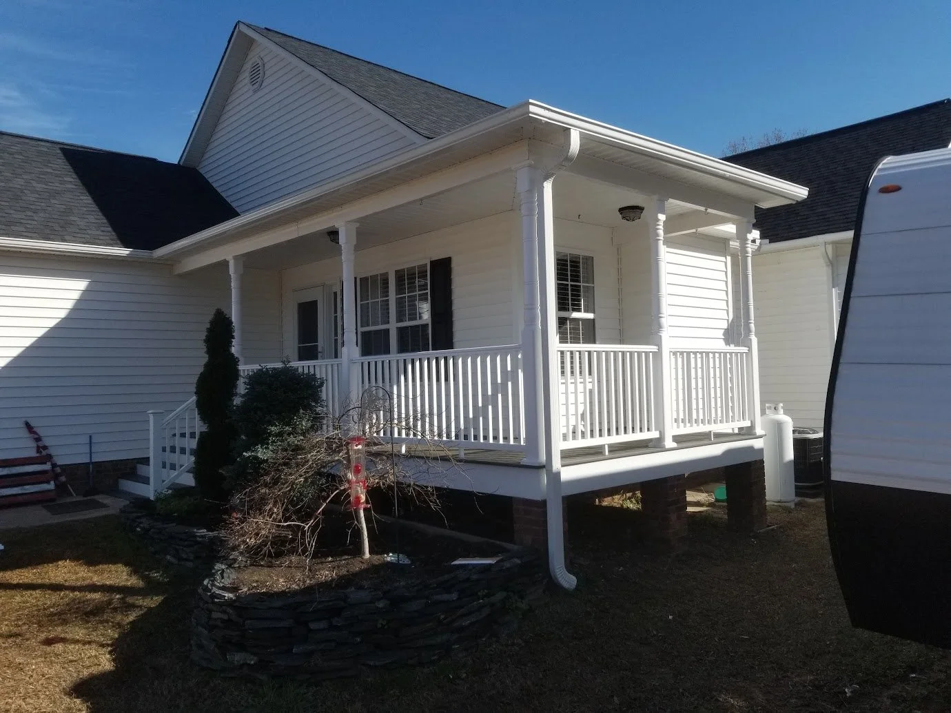 White house with a front porch, black shutters, and a small garden bed with a shrub and leafless tree in front, during daytime.